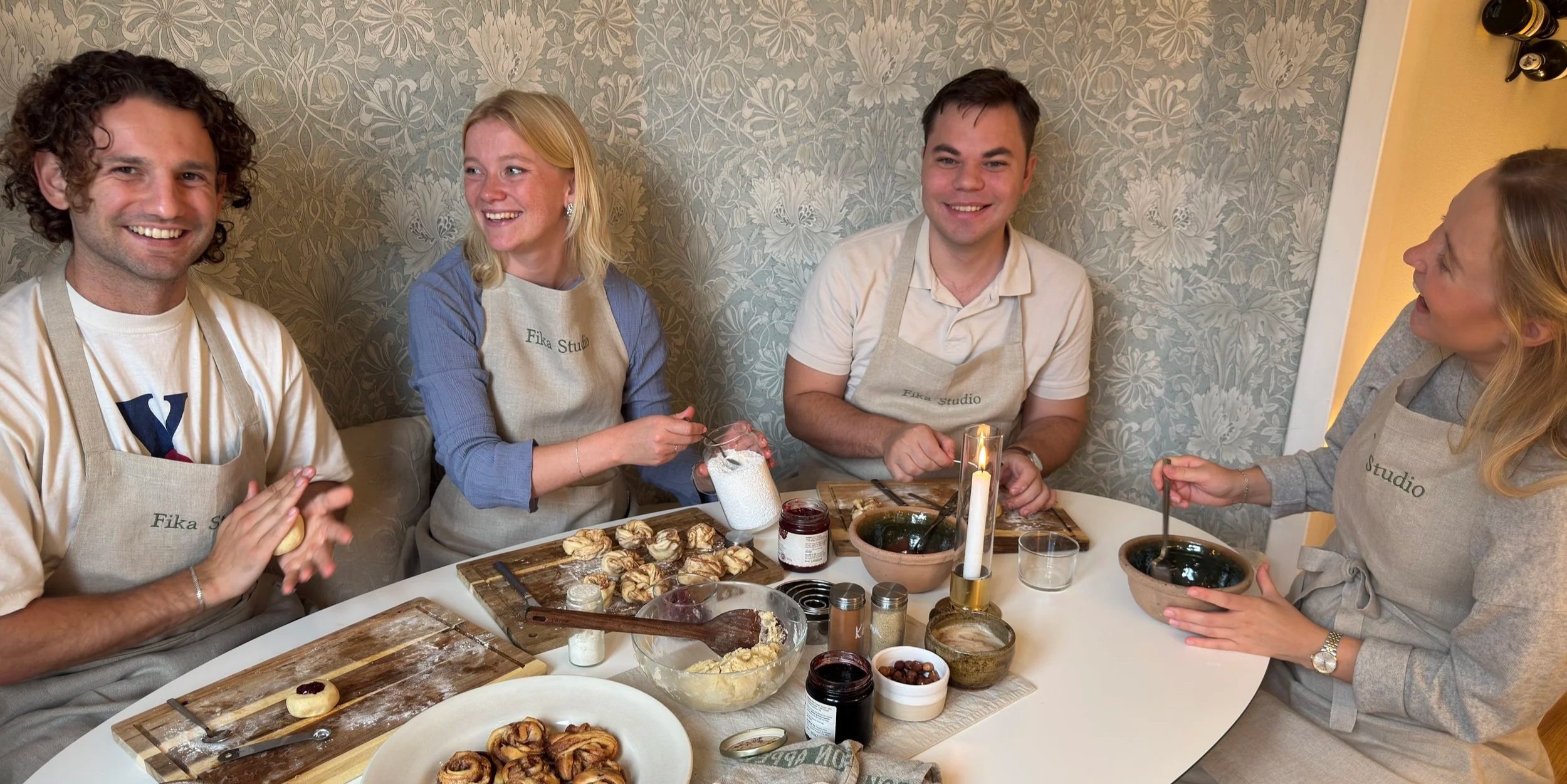 Five people wearing aprons with "Fika Studio" on them sitting around a table with baked goods, bowls, and utensils, smiling and enjoying a baking activity in a cozy room with floral wallpaper.