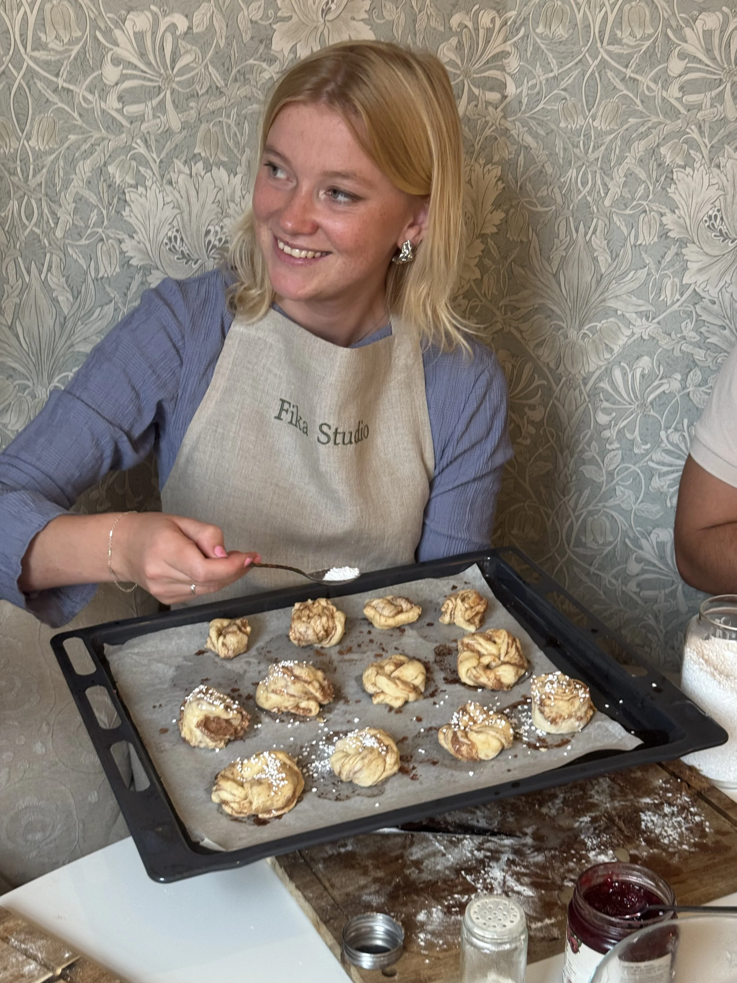 A woman wearing a beige apron with 'Fika Studio' written on it is smiling while holding a spoonful of powdered sugar over a baking tray filled with freshly baked cinnamon rolls. The tray is on a wooden table with various jars and a white dish nearby.