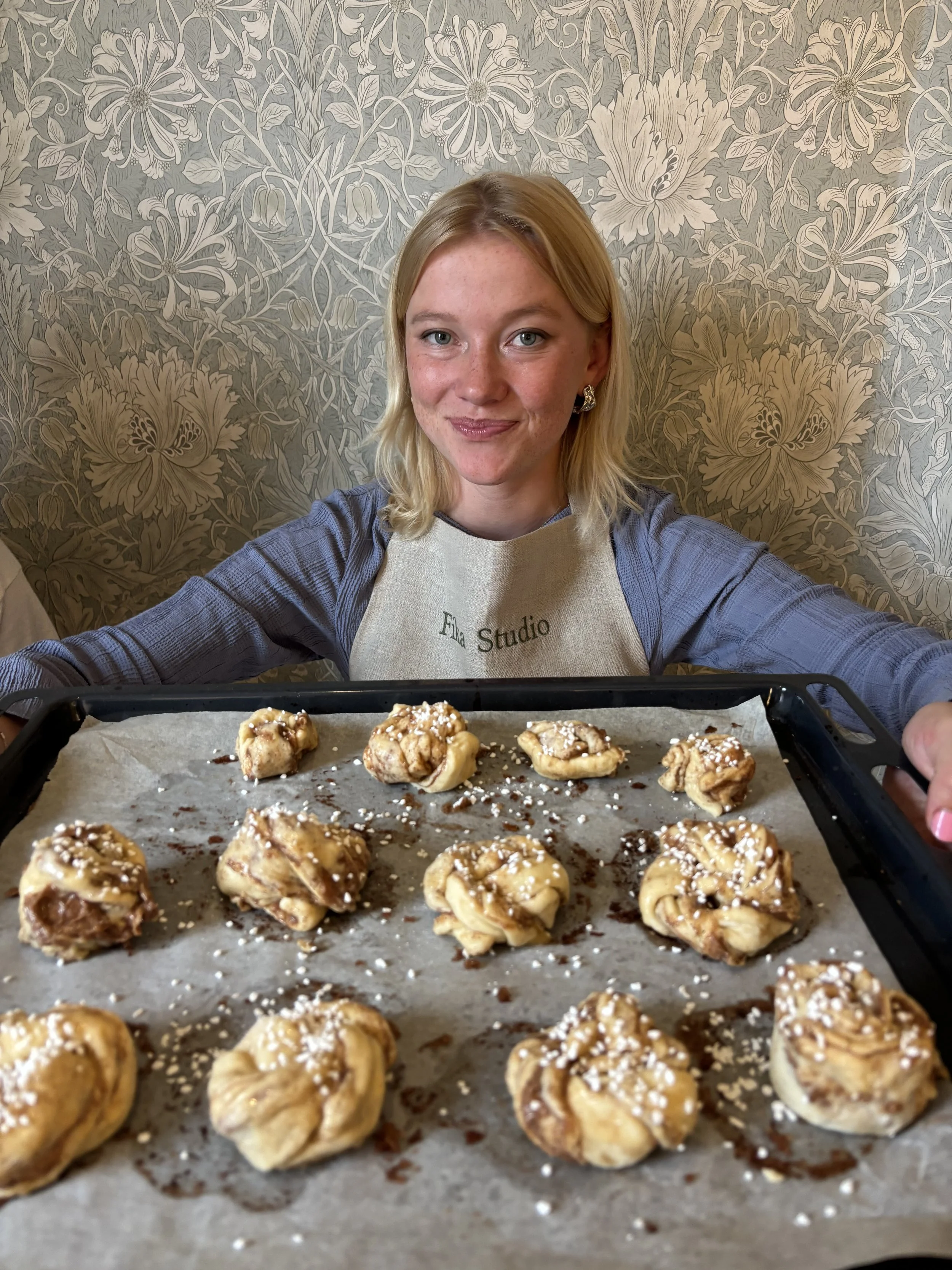 A woman wearing an apron with the text 'Ella Studio' and a striped shirt is smiling at the camera, holding a tray of freshly baked cinnamon rolls topped with icing and sprinkles.