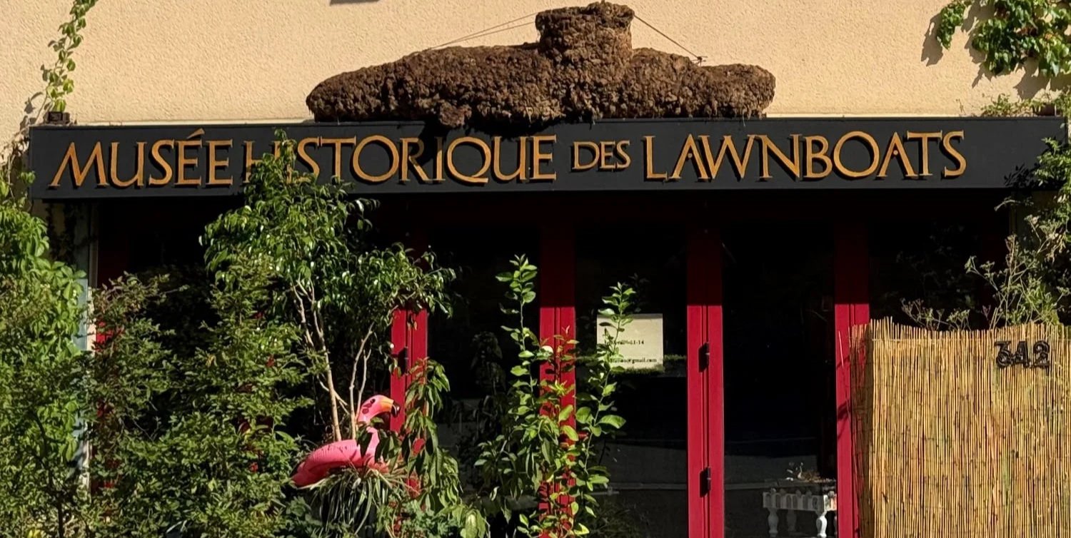 Entrée du musée historique des yachts avec un panneau noir à lettres dorées, entourée de plantes et un flamant rose en plastique.