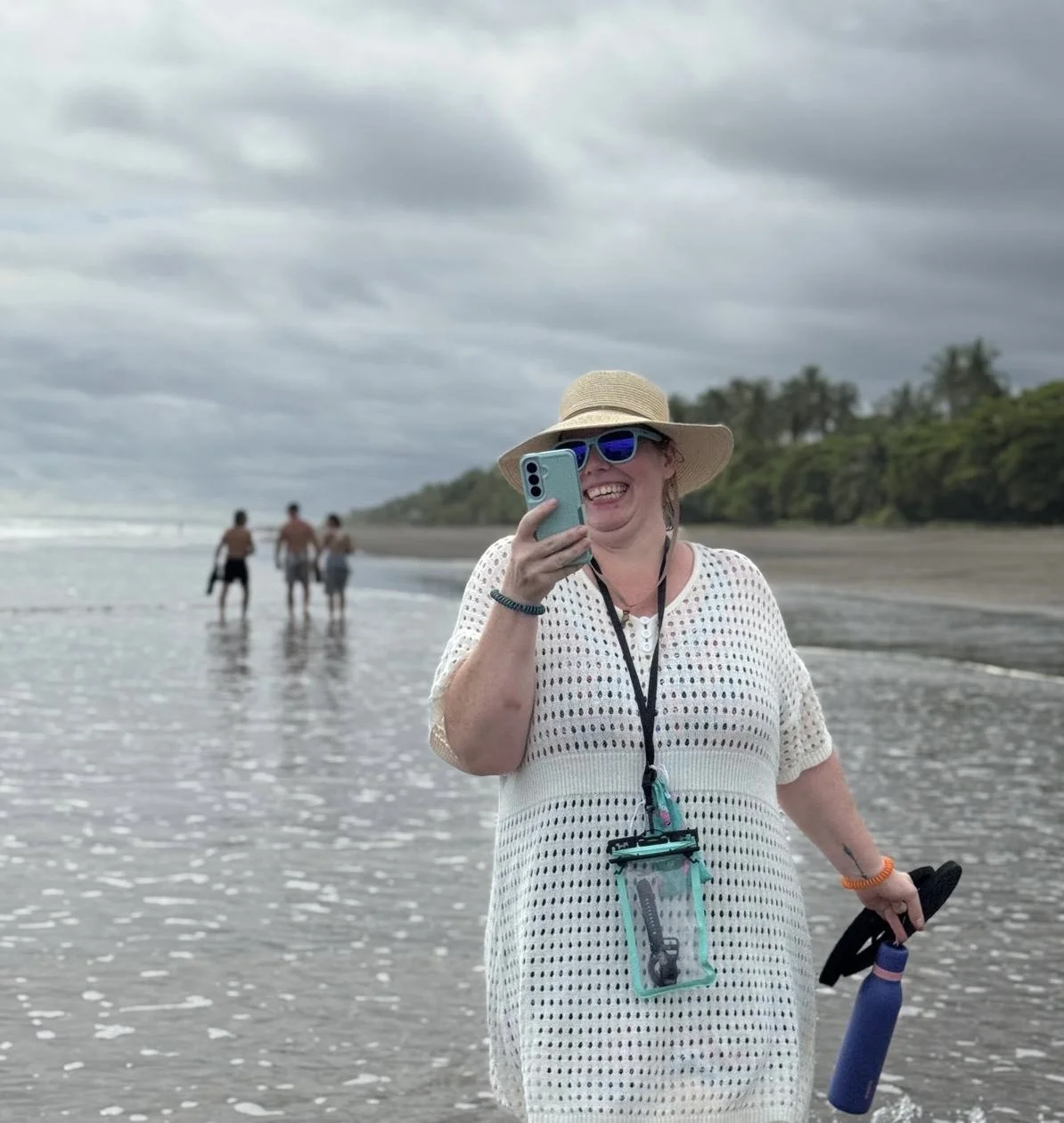 A woman smiling and taking a photo with her phone on a beach with others in the background, overcast skies, wearing a sun hat, sunglasses, a white crochet dress, and holding a water bottle.