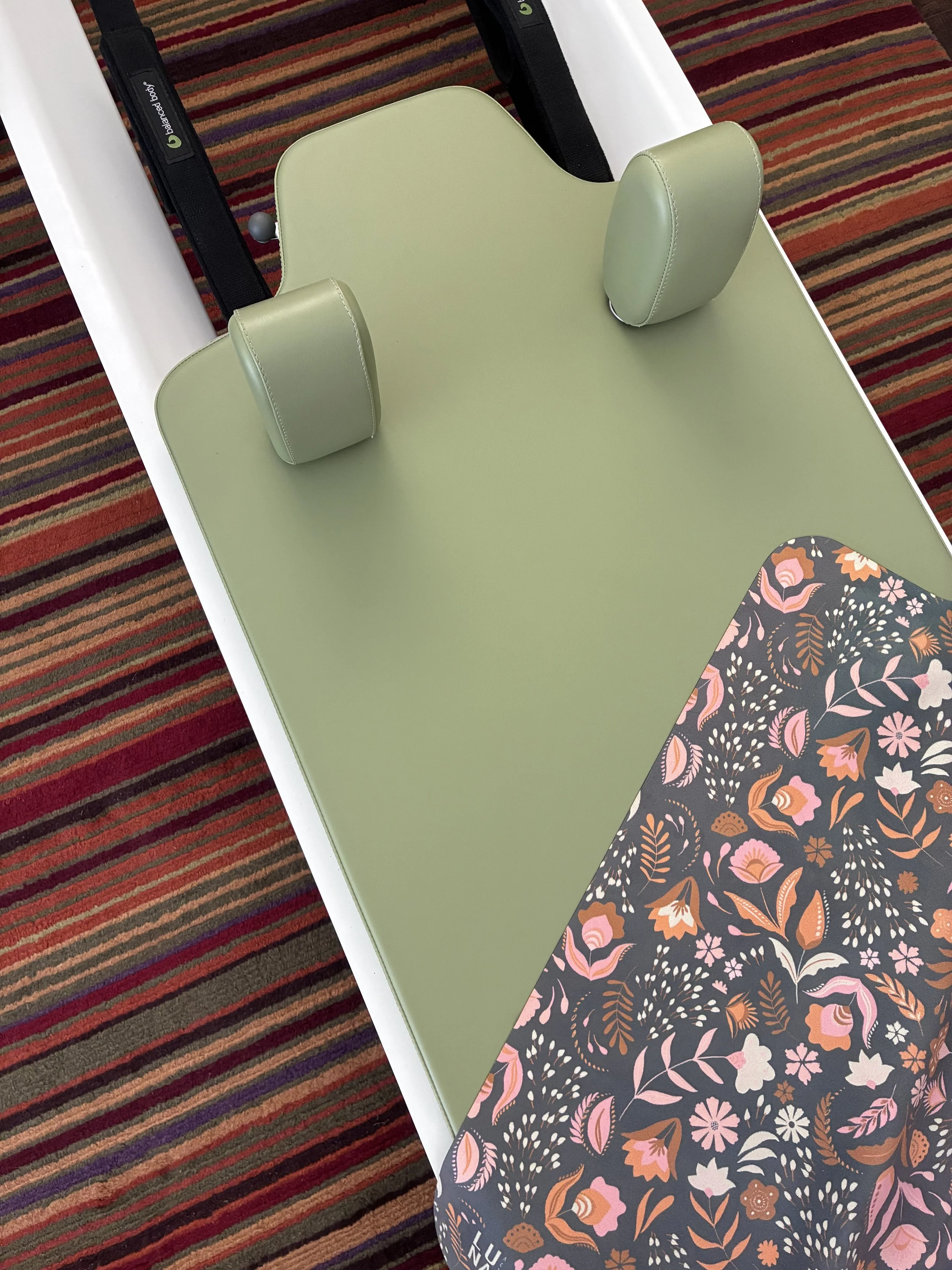 A green Pilates Reformer carriage on white rails, with a patterned fabric grippy cover, and a colorful striped carpet.