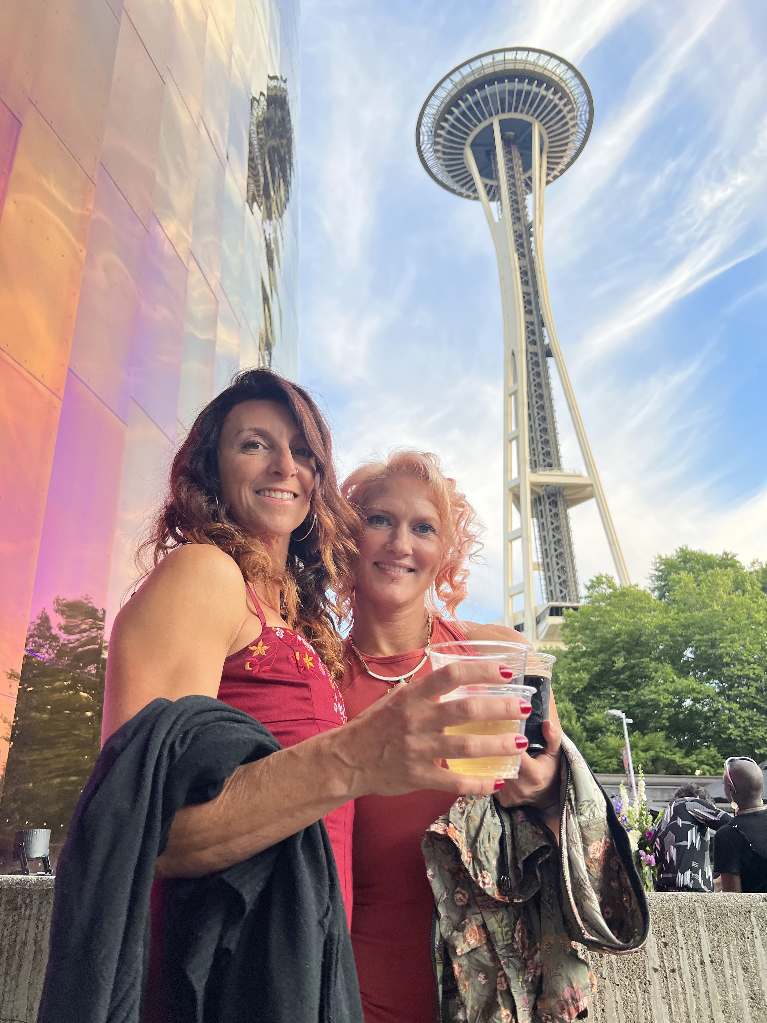 Two women smiling at the camera while holding drinks outdoors with the Space Needle in Seattle, Washington, in the background.