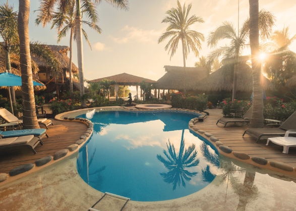 Resort pool area with palm trees, lounge chairs, and a thatched-roof hut at sunset.