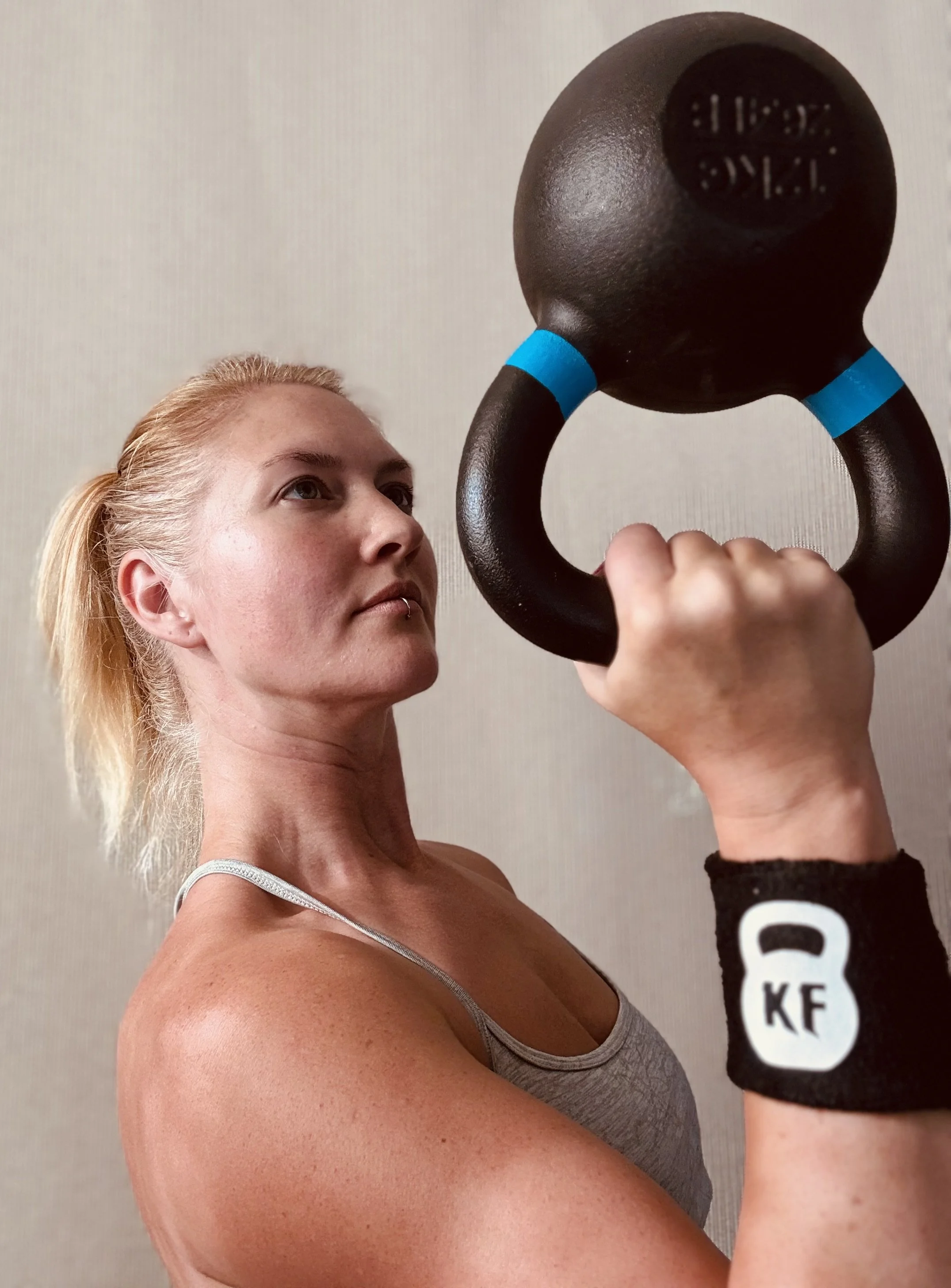 A woman with blonde hair lifting a black kettlebell upside down, looking up at it.