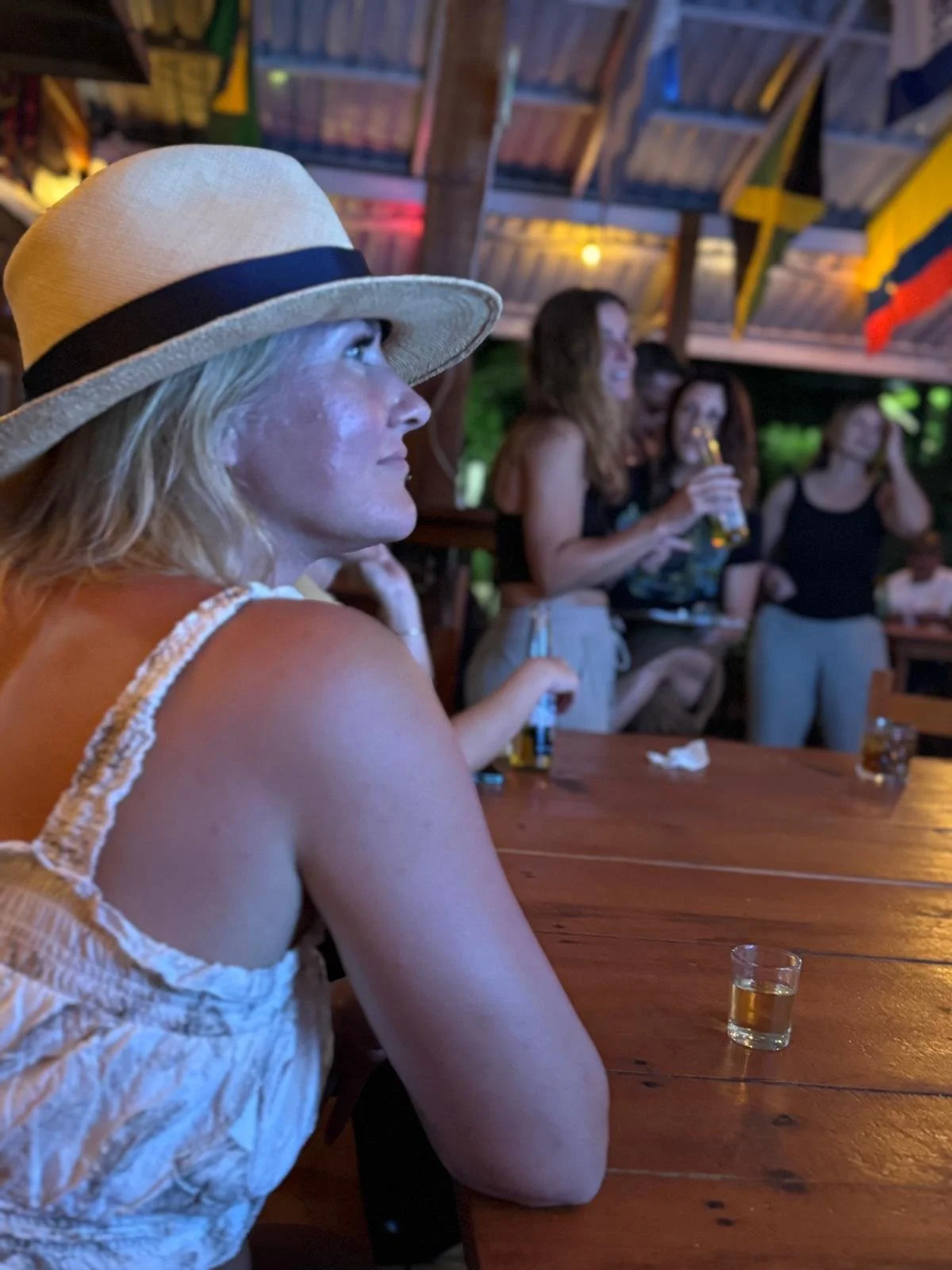 A woman with blonde hair wearing a straw hat and a sleeveless top is sitting at a wooden table, looking to the side. In the background, there are several women standing and singing karaoke, holding drinks and smiling.