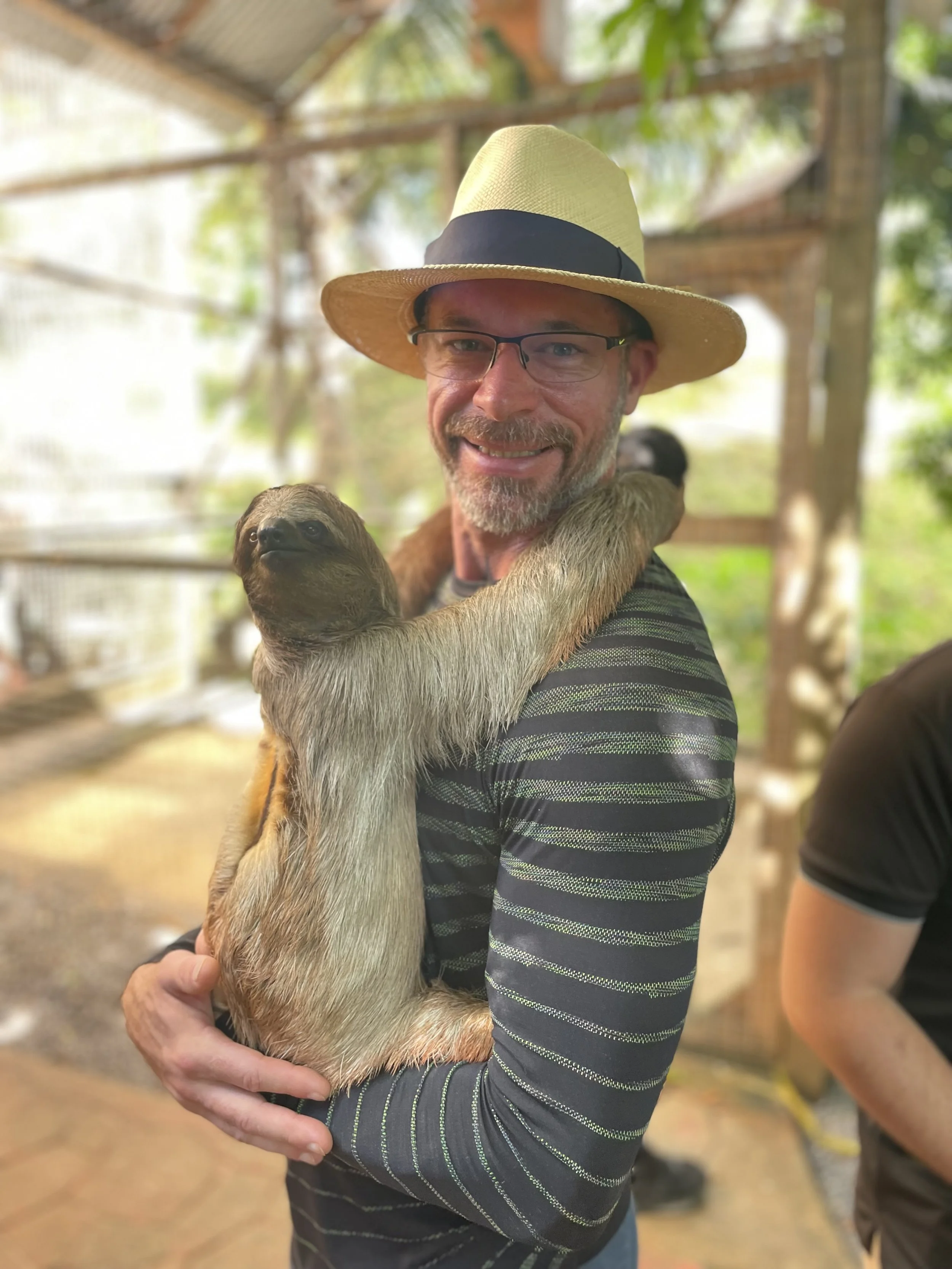 A smiling man wearing glasses and a straw hat, holding a sloth with its arms around his neck inside a shaded enclosure with  trees in the background.
