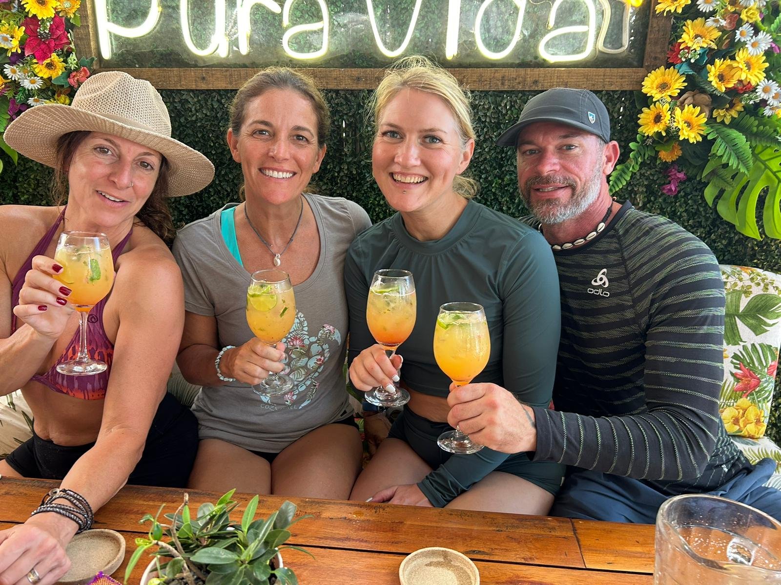Four people sitting together at a table, holding colorful drinks, smiling, with a floral and greenery background and a sign that says 'Pura Vida'.