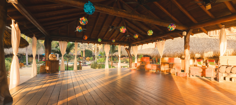 Open-air wooden pavilion decorated with colorful paper lanterns, with cushioned seating and drapery, in a tropical setting with thatched structures and lush greenery.