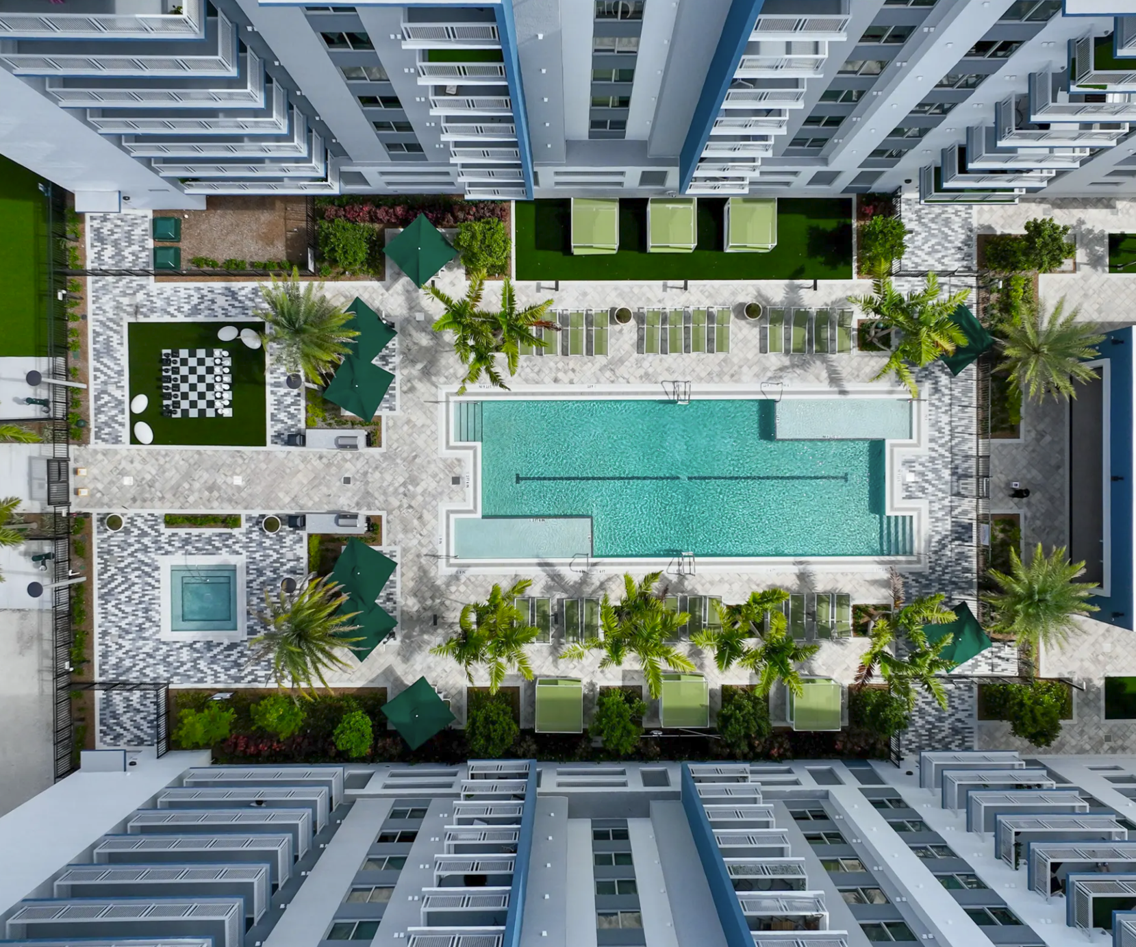 Aerial view of a modern apartment complex courtyard featuring a swimming pool, green lawn, lounge chairs, umbrellas, palm trees, and outdoor seating areas surrounded by tall apartment buildings.