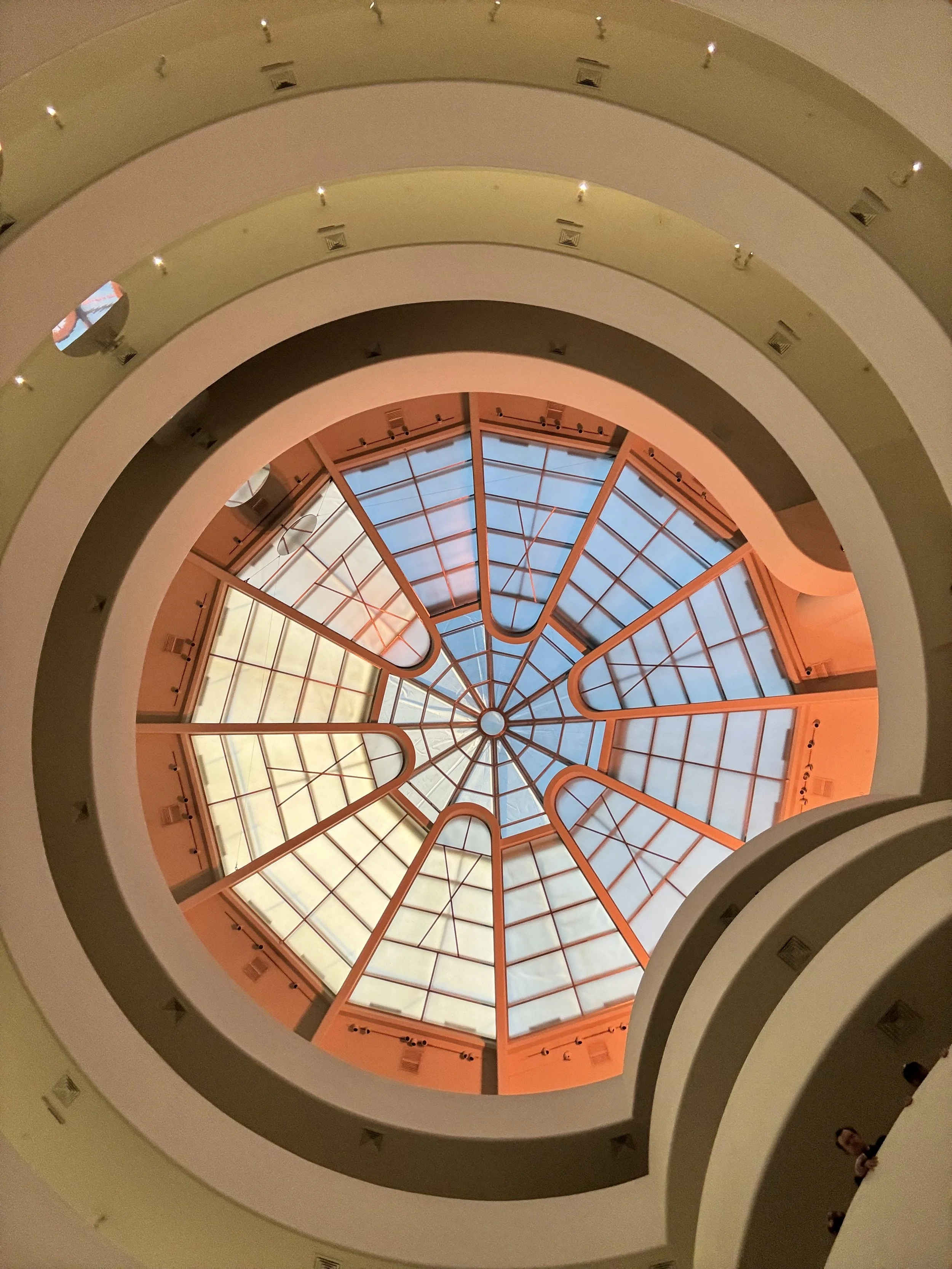 View looking up through multiple levels of a circular interior with a glass-domed ceiling at the top, framed by curved balconies painted in cream and yellow shades.