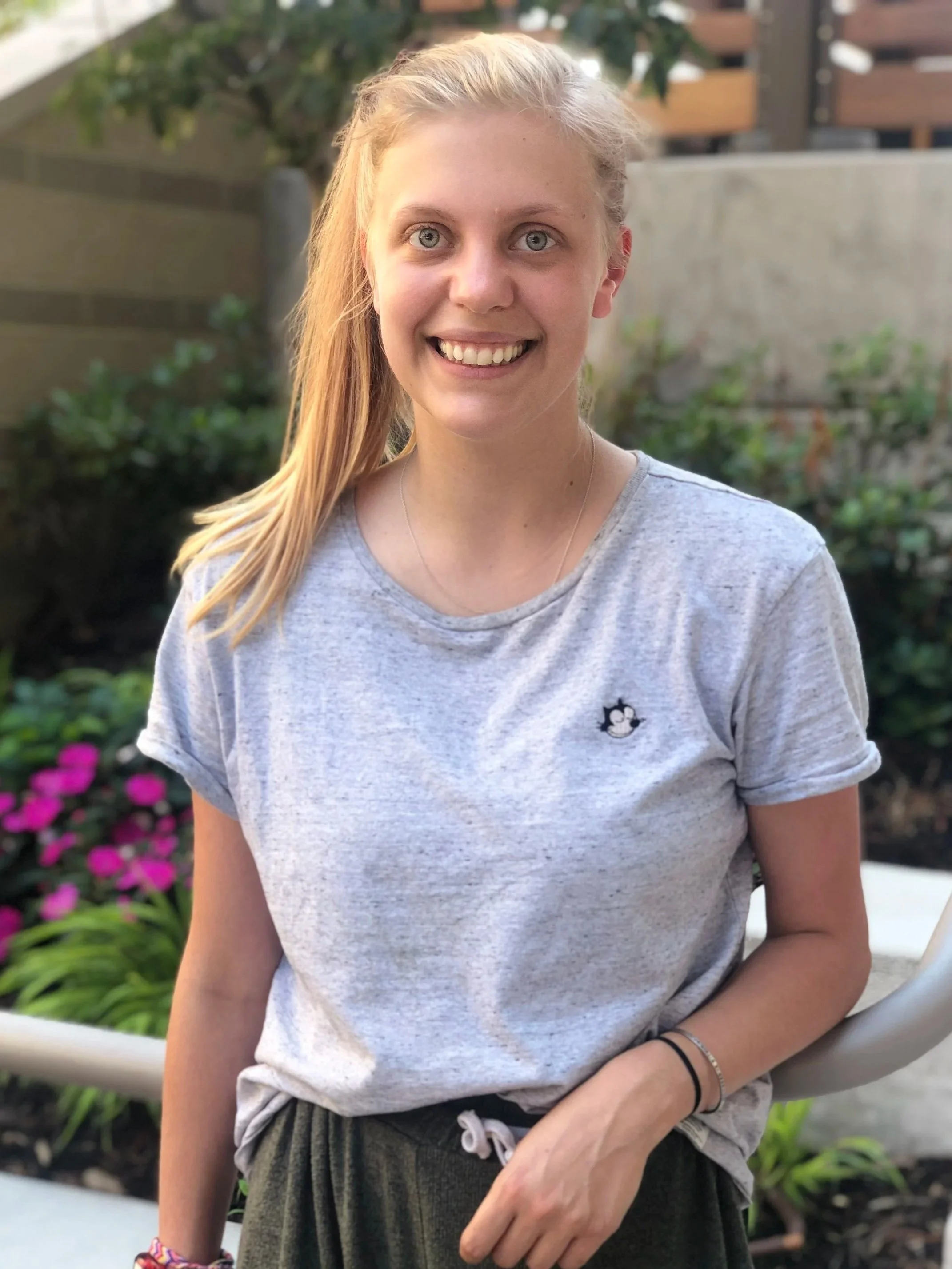 Close-up of a young woman with blonde hair in a ponytail, smiling outdoors near a garden with pink flowers and greenery, wearing a light gray t-shirt.