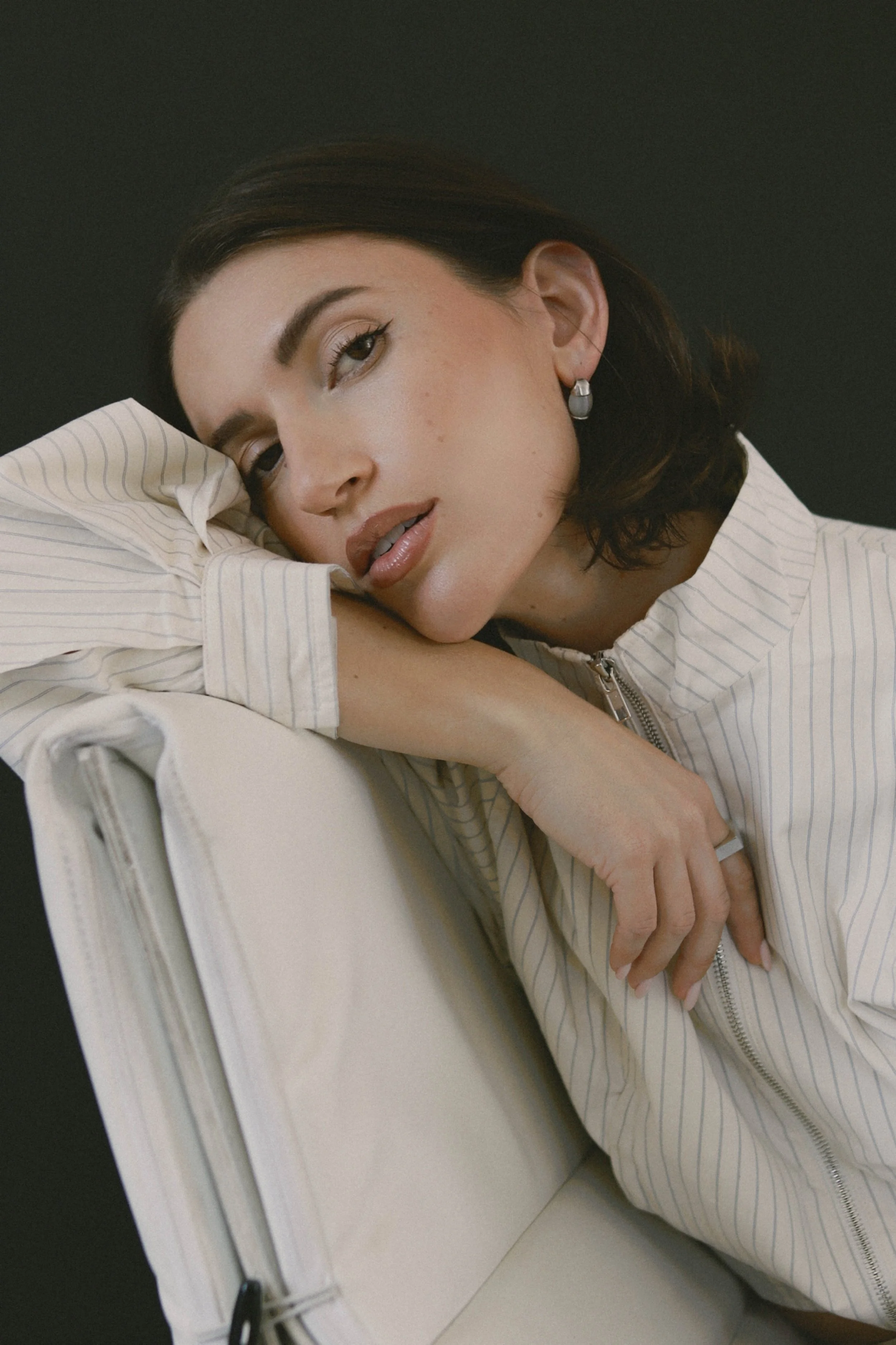 A woman with dark hair, wearing earrings and a striped zip-up jacket, is resting her head on her arm on a white surface with a black background.