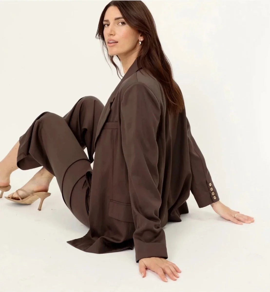 Woman in brown suit sitting on ground, with one knee up and one hand on the ground, wearing high heels and earrings, with long brown hair and a neutral expression.