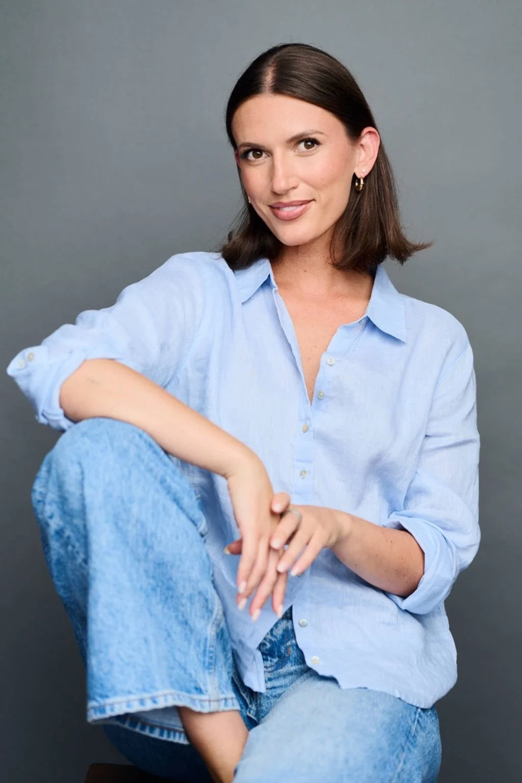 A woman with shoulder-length brown hair, wearing a light blue button-up shirt and blue jeans, sitting against a gray background, smiling and looking at the camera.