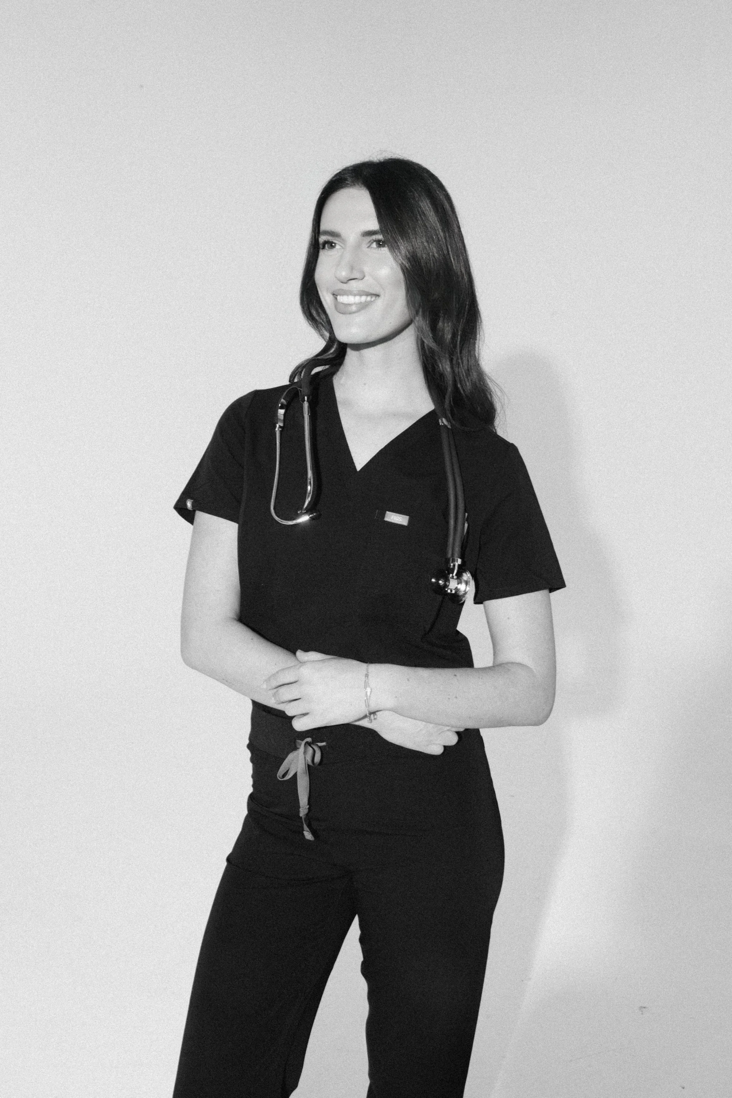 Black and white photo of a female healthcare professional wearing scrubs with a stethoscope around her neck, standing against a plain background.