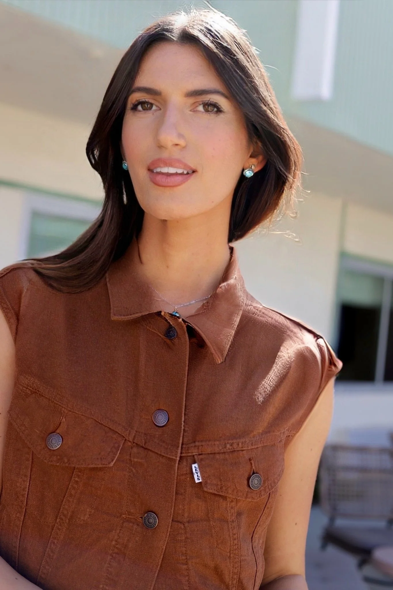 A woman with long dark hair and earrings smiling outdoors, wearing a brown sleeveless button-up shirt.