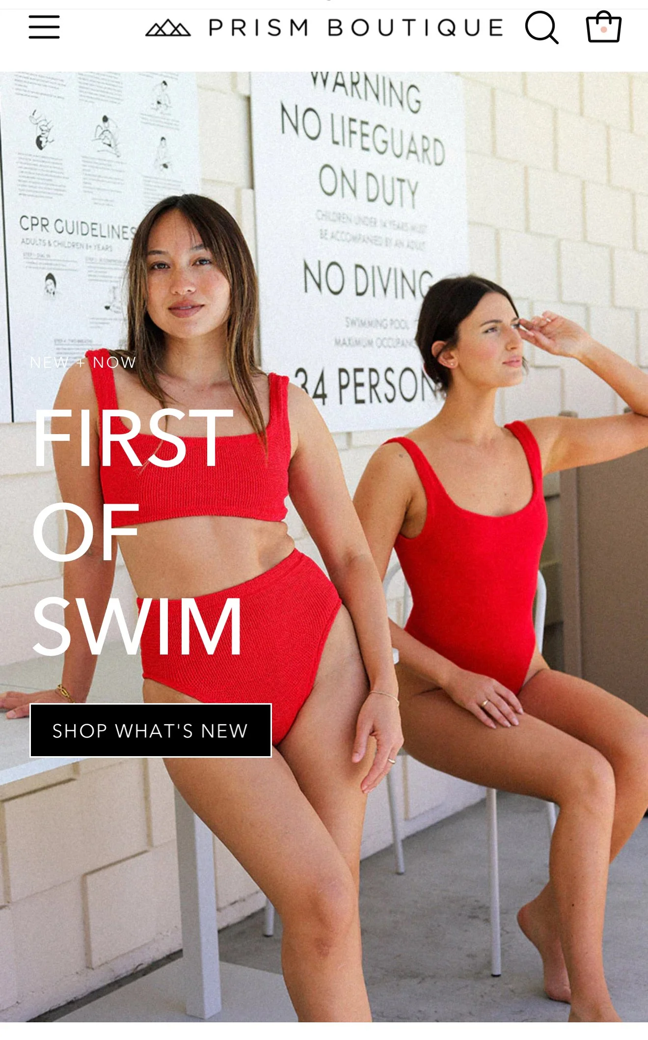Two women in red swimsuits sitting against a white brick wall, with swim-related informational posters hanging behind them, on a gray concrete floor.