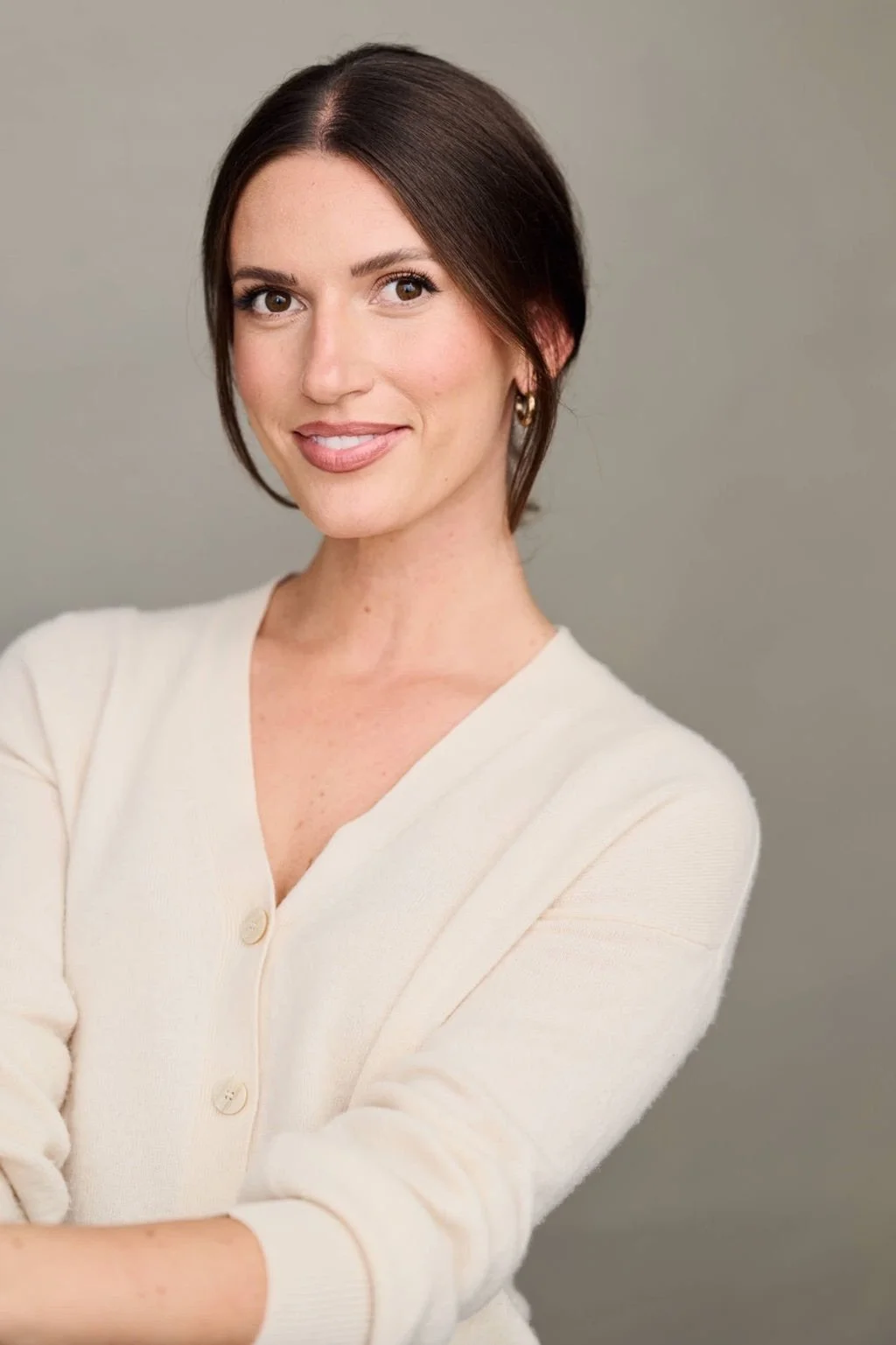 A woman with dark brown hair and fair skin, smiling, wearing a light-colored cardigan with a V-neck, standing against a plain gray background.