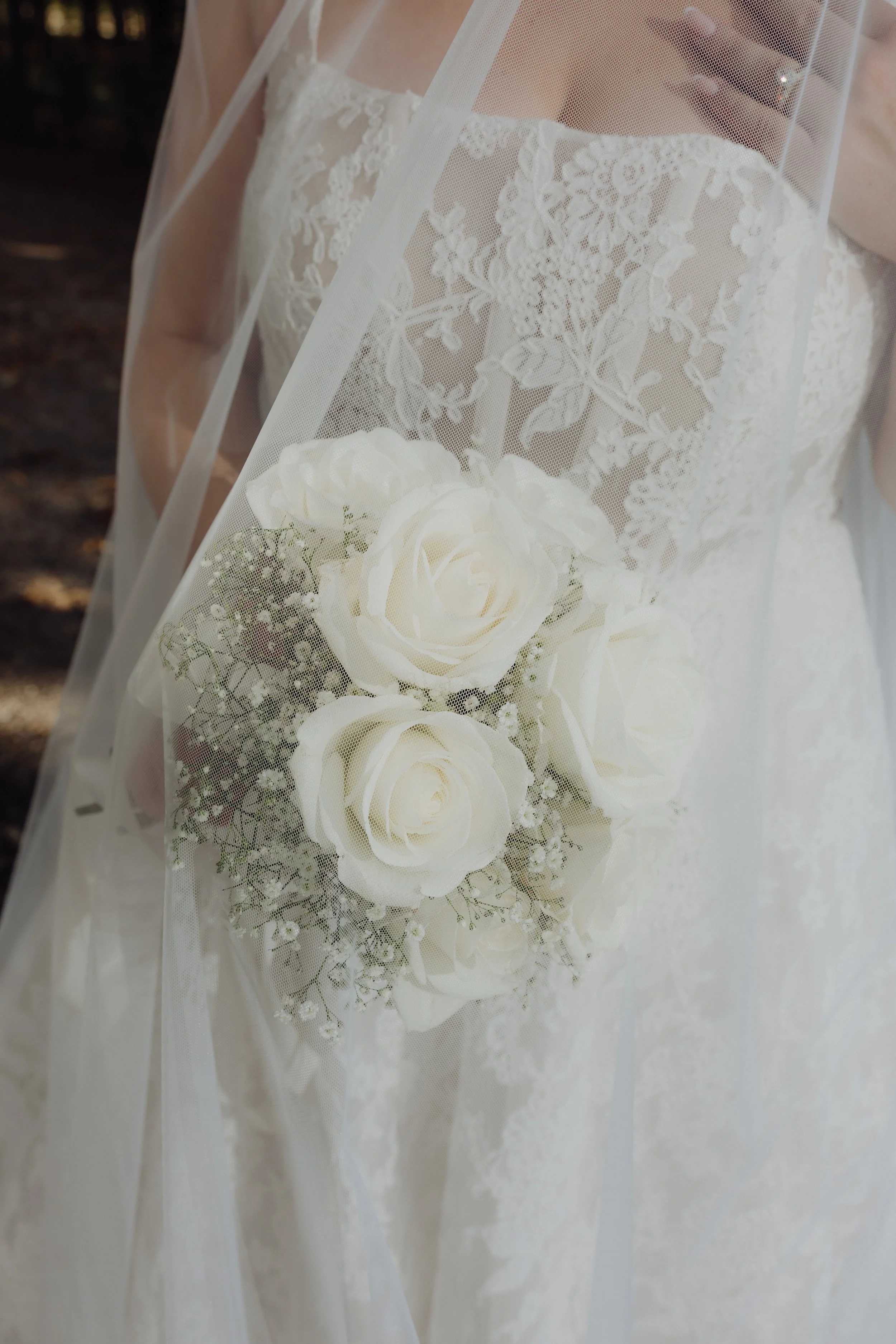 Elegant bridal bouquet of white roses resting against a delicate lace wedding dress and veil.