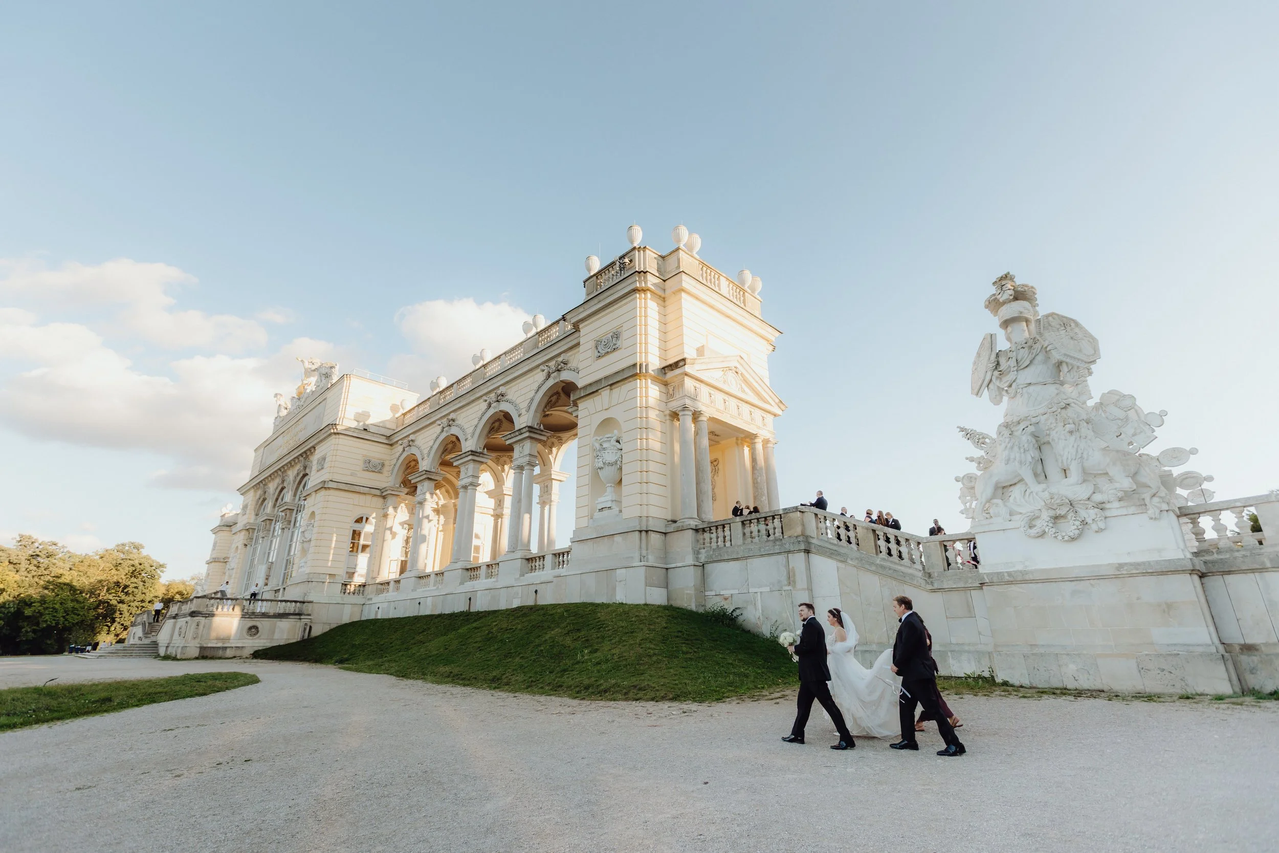 Bride and groom walking with a wedding attendant in front of the historic Gloriette at Schönbrunn Palace in Vienna, Austria.