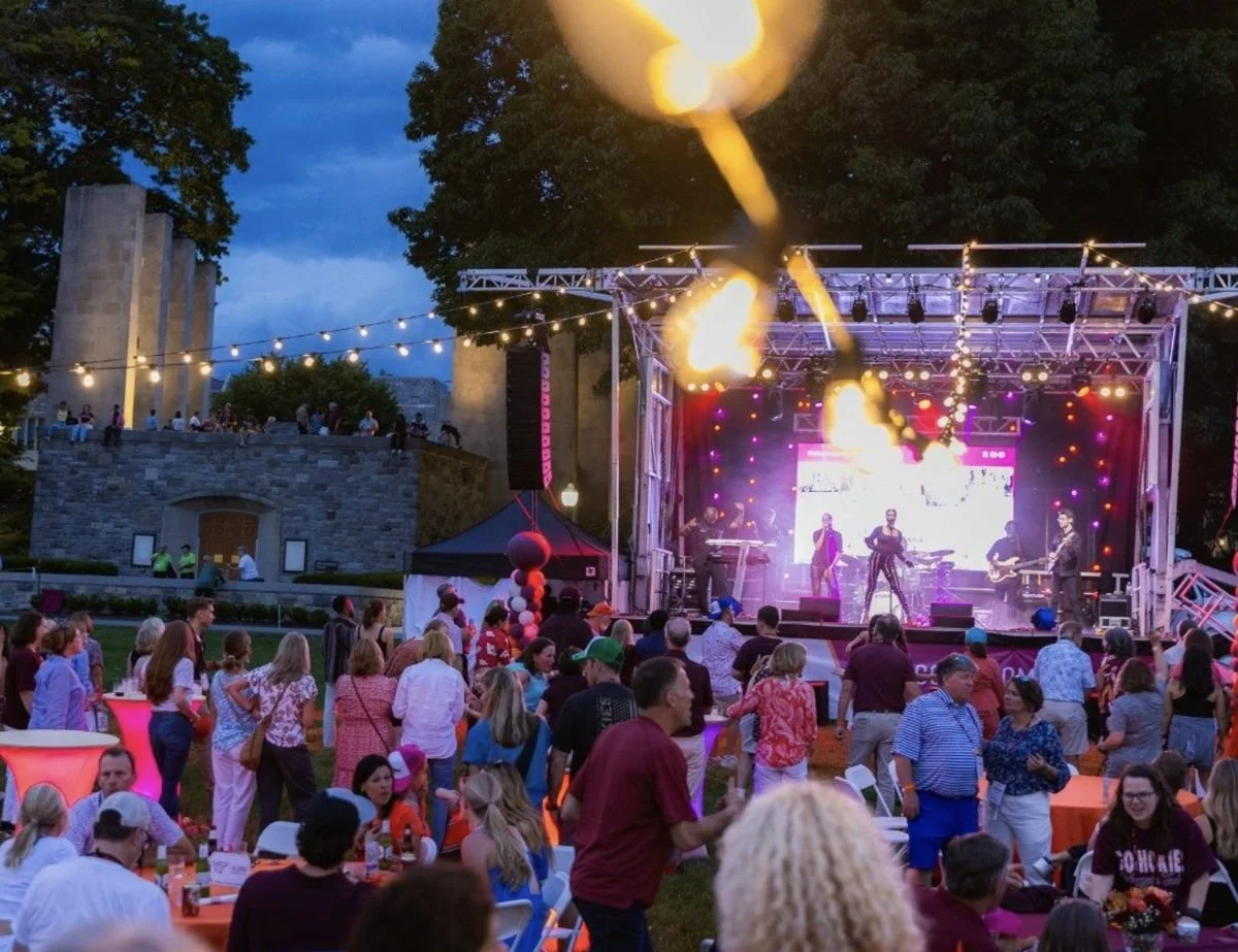 Alumni gather at dusk in front of an outdoor concert stage with string lights and fire effects during Alumni Weekend.