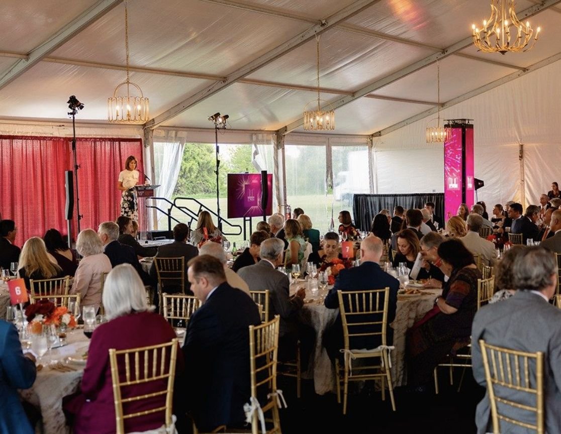 Guests seated at round tables in a chandelier-lit tent listen to a speaker at the Ut Prosim Society Weekend dinner.