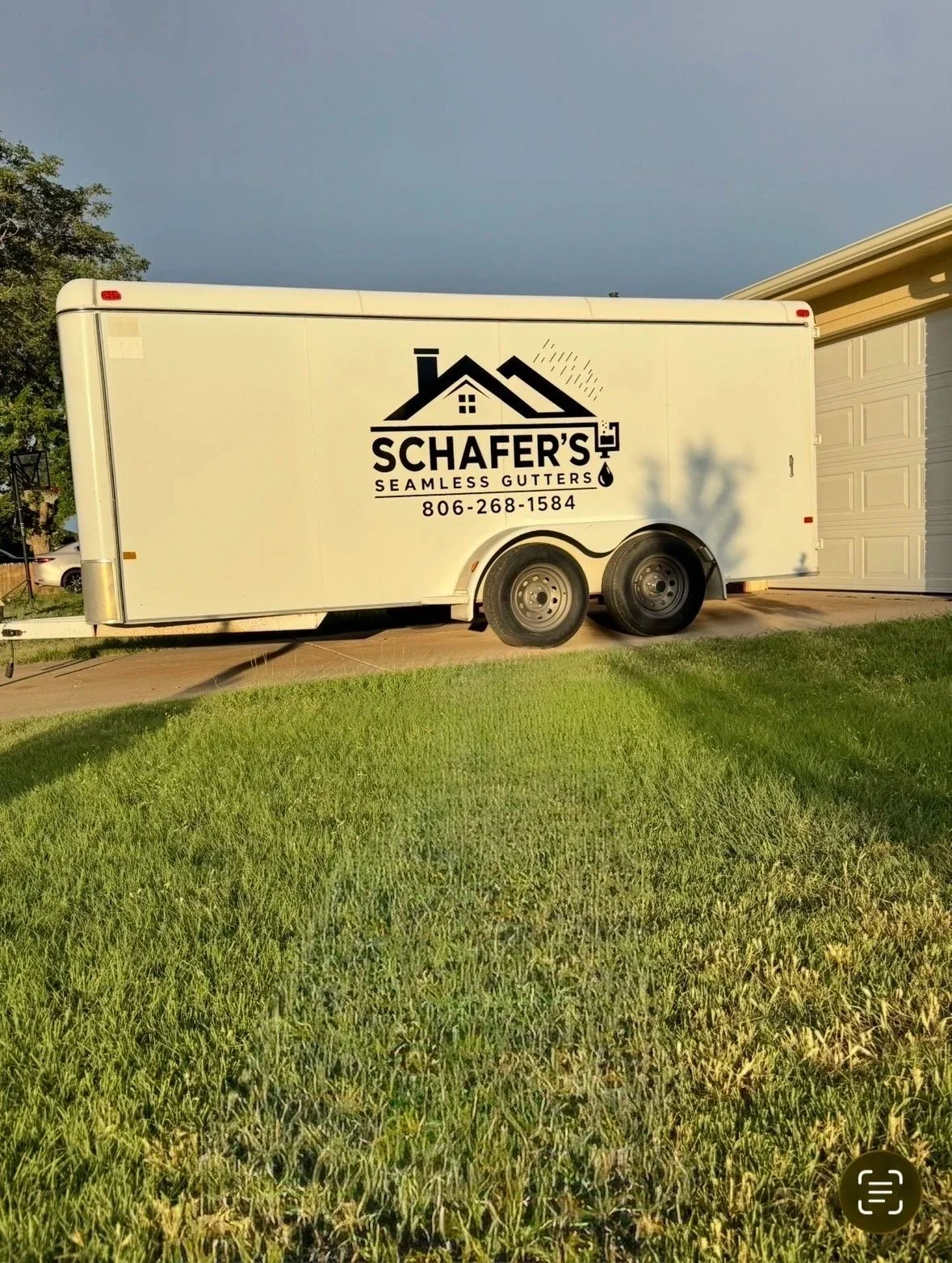 A white trailer with the logo and contact information for Schafer's Seamless Gutters parked on a driveway next to a house, with a green lawn in the foreground and a cloudy sky above.