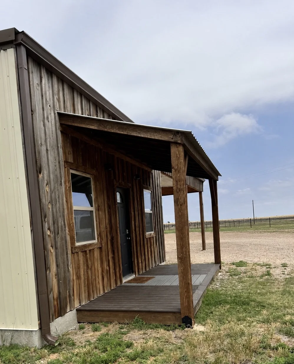 A small wooden porch attached to a rustic barn with weathered wood siding, located in a rural area with grass and dirt in the surroundings.
