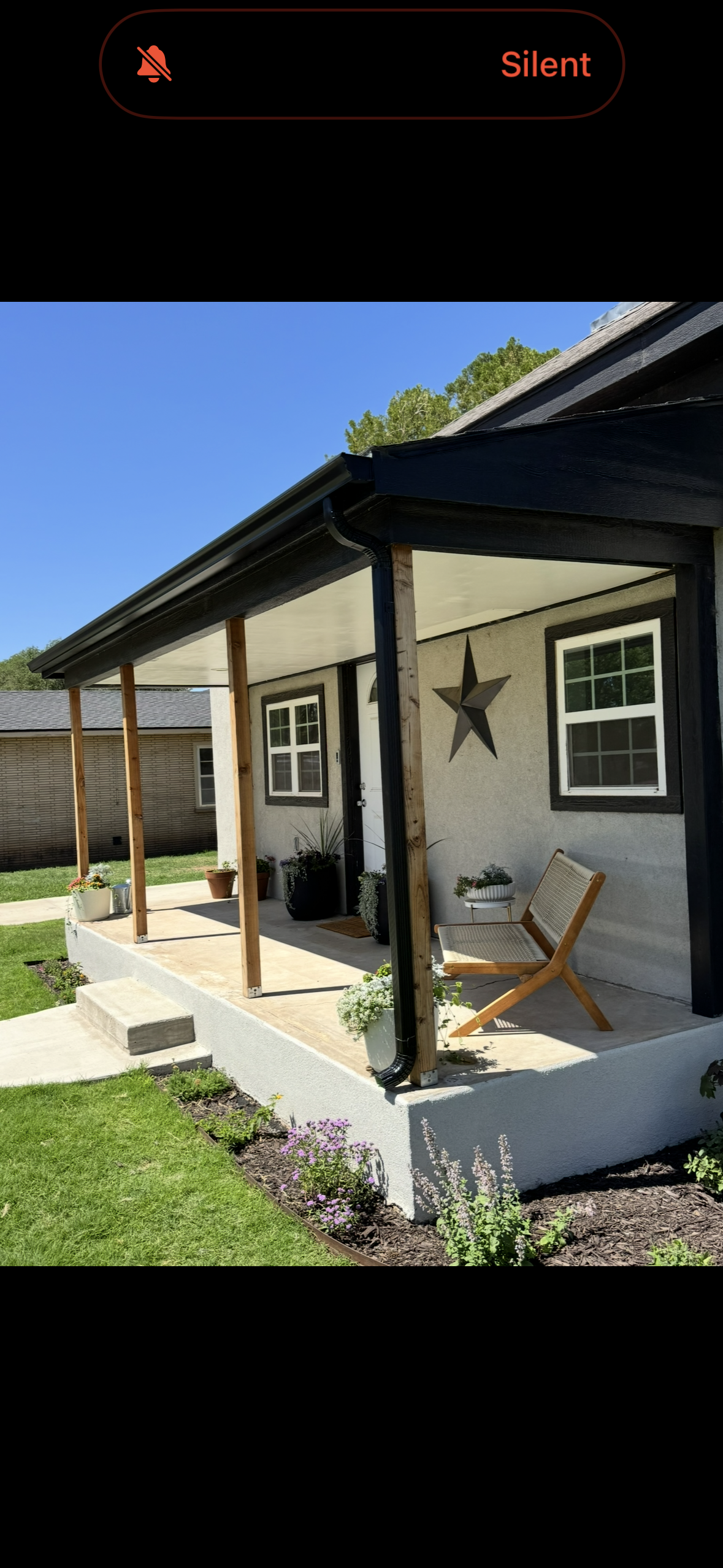 Front porch with potted plants, a wooden bench, and a star wall decor on a house with a gray stucco exterior and black trim, under a blue sky.