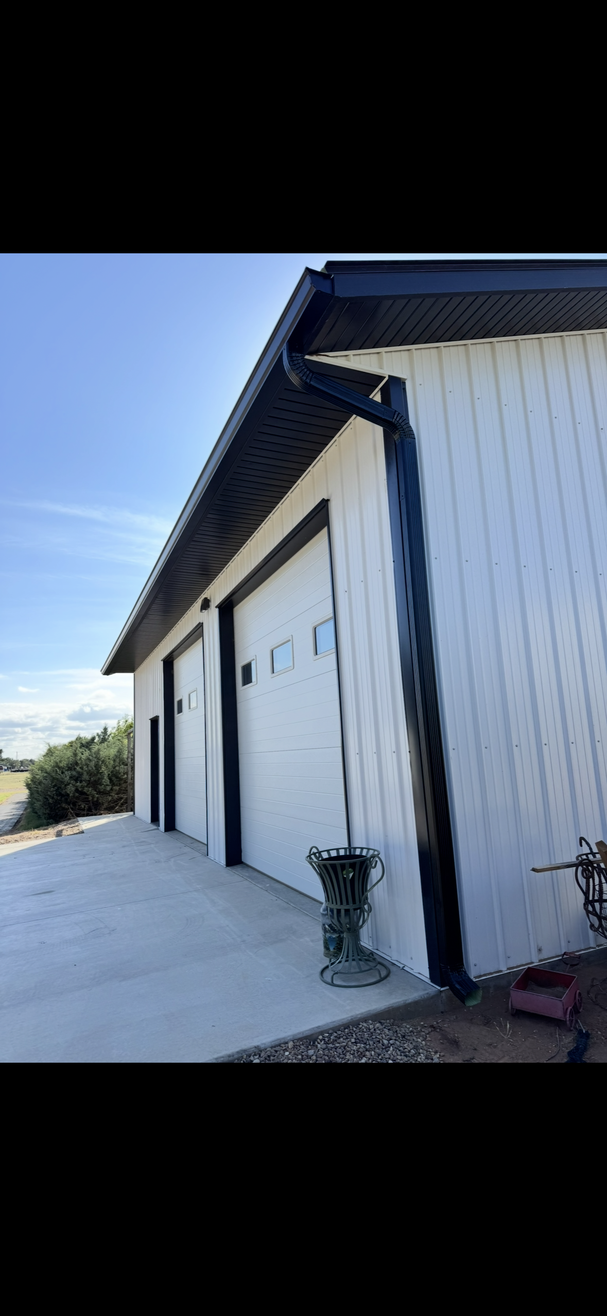 Metal building with four garage doors, black gutters, and a concrete driveway, with a blue sky and some bushes in the background.