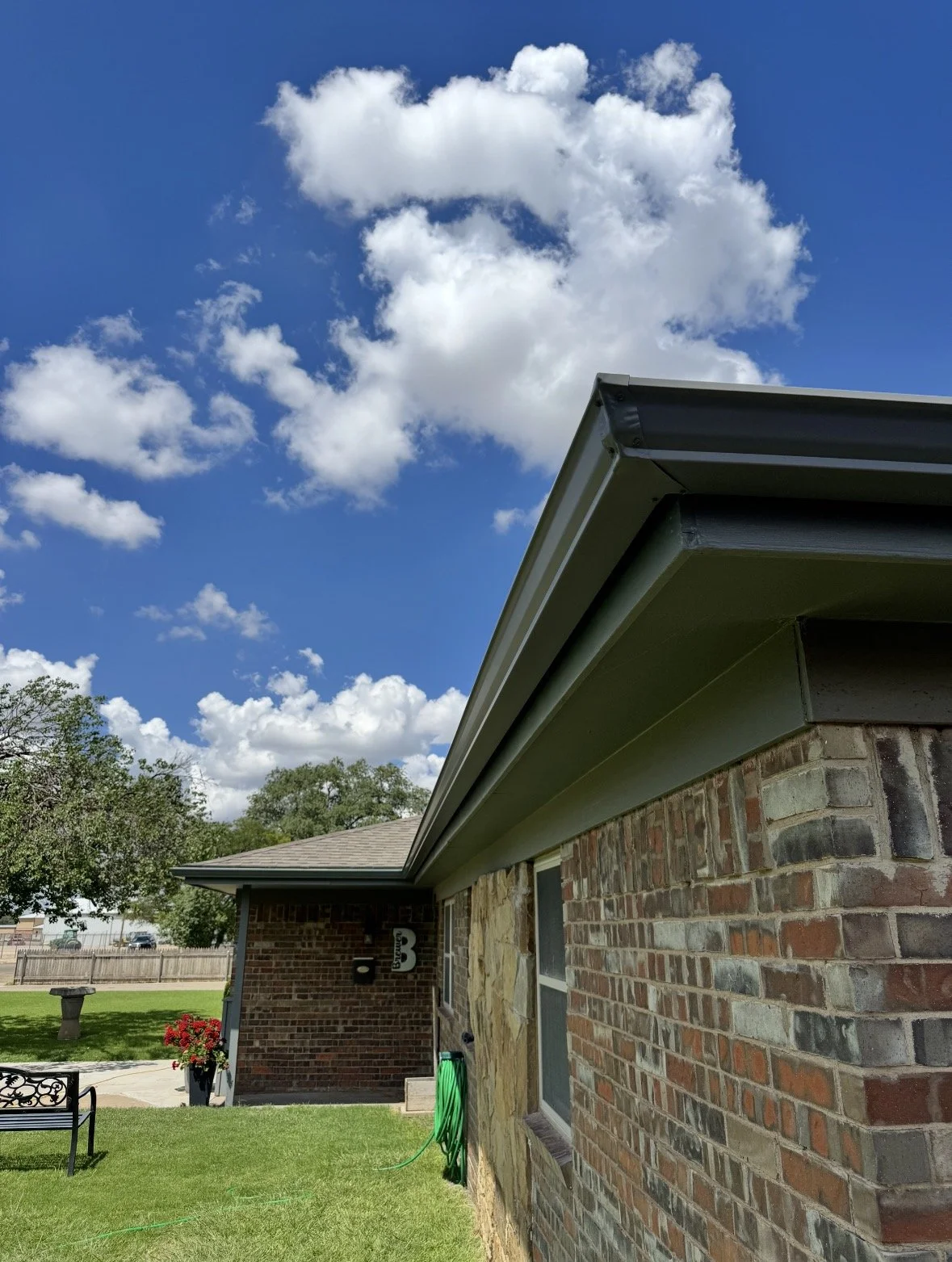 Side view of a brick house with a black gutter, green garden hose, and a backyard with trees, a bench, and a trash bin under a blue sky with scattered clouds.