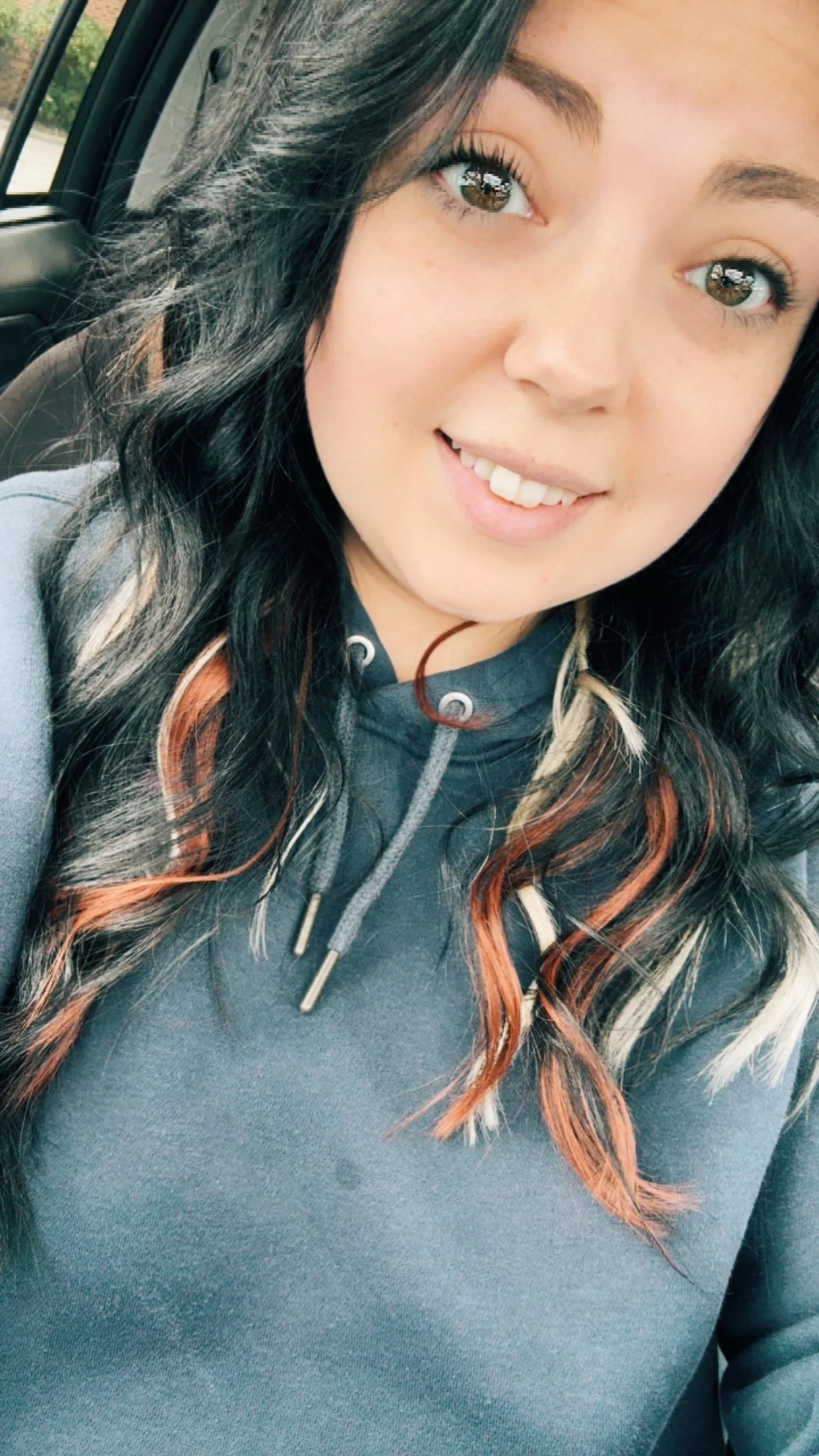 Close-up of a smiling young woman with long black hair with orange and blonde streaks, wearing a gray hoodie, inside a vehicle.