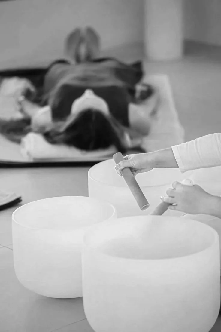 A person uses a mallet to strike a singing bowl while a dog lies on a bed in the background.