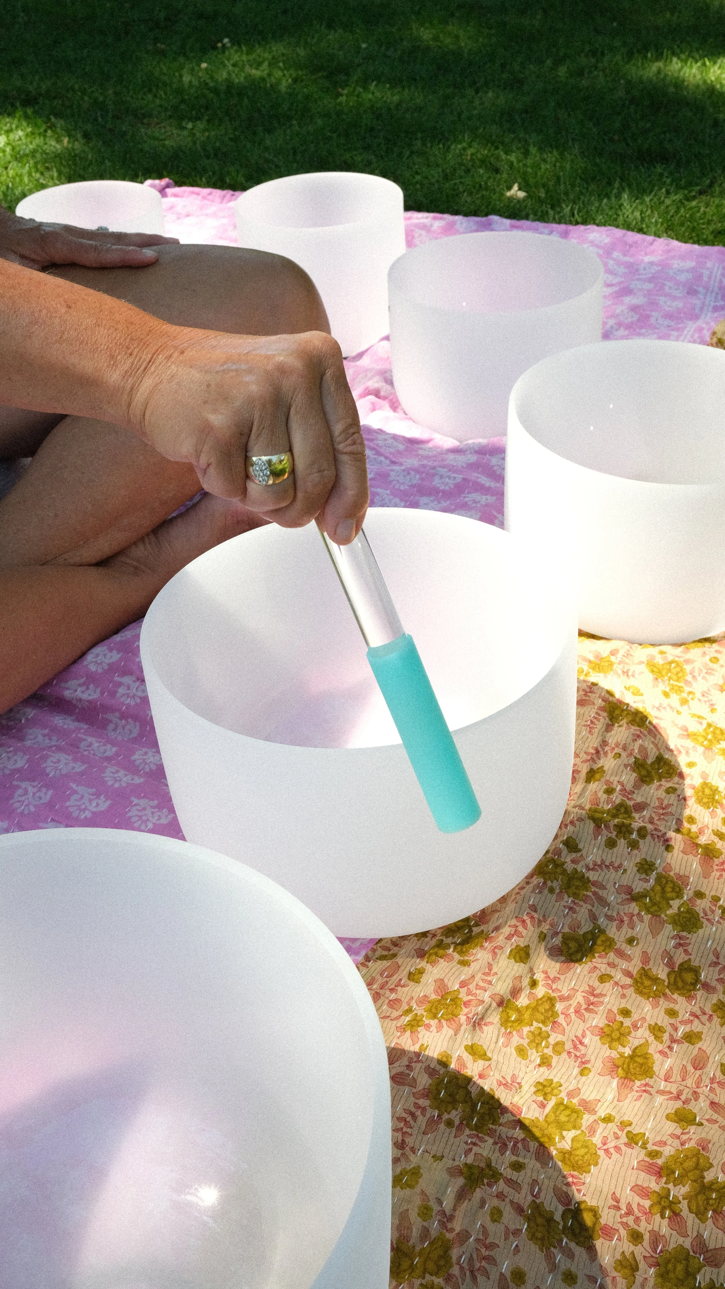 Person playing singing bowls outdoors on a pink patterned fabric with green grass in the background.