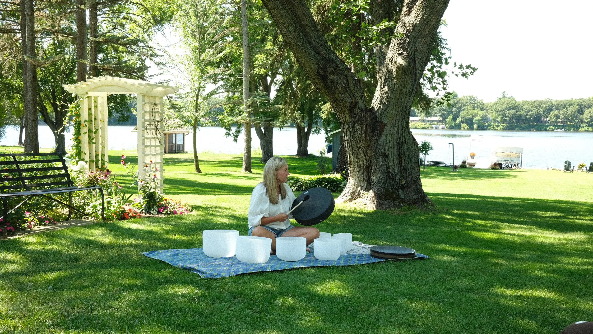 A woman sitting outdoors on a grassy area near a lake, holding a black pan, with singing bowls and a mallet on a blanket, surrounded by trees, flowers, and outdoor furniture.