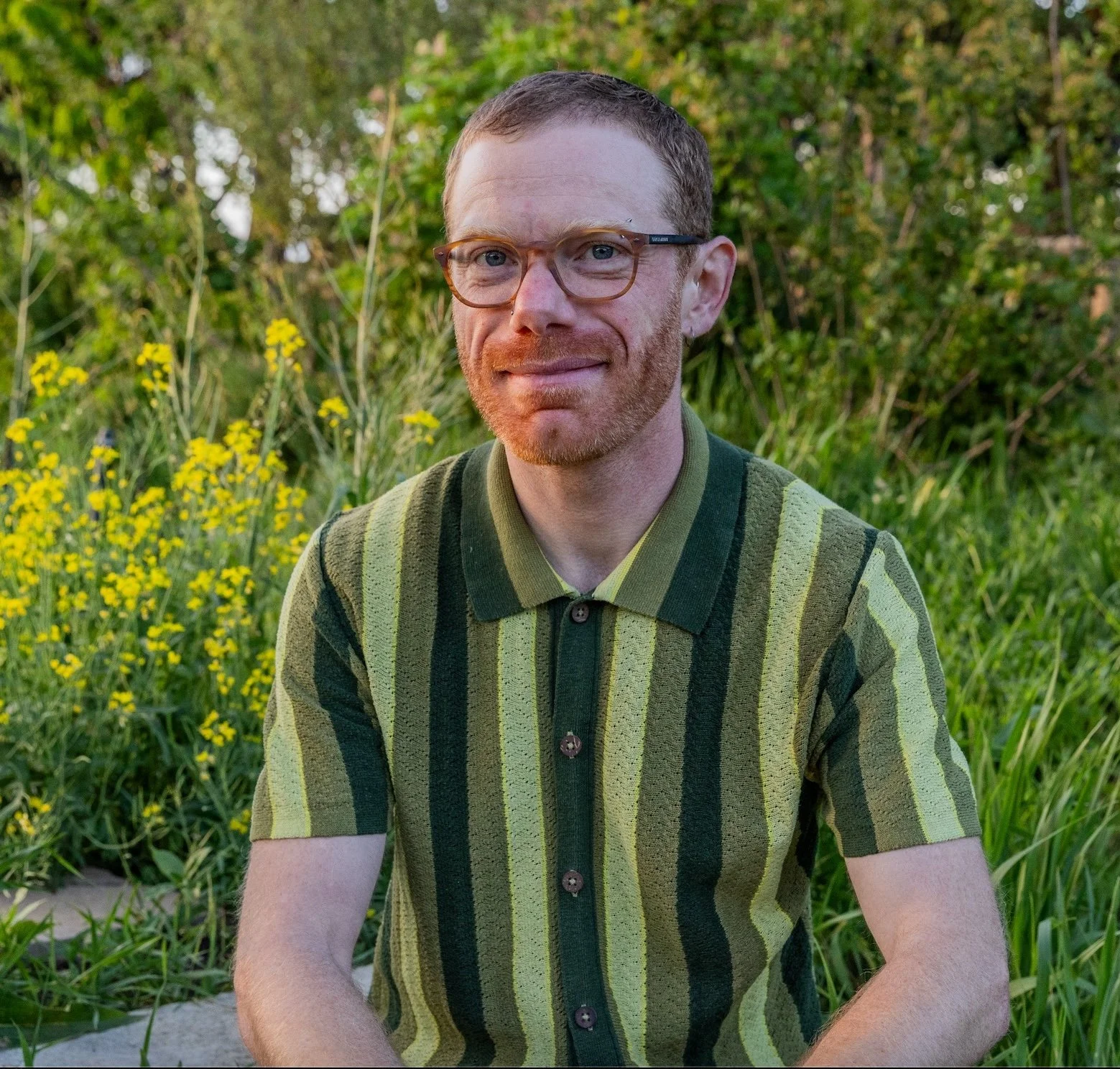 A man with glasses and a beard sitting outdoors on a grassy area with yellow flowers and green trees in the background, wearing a striped green short-sleeve polo shirt.
