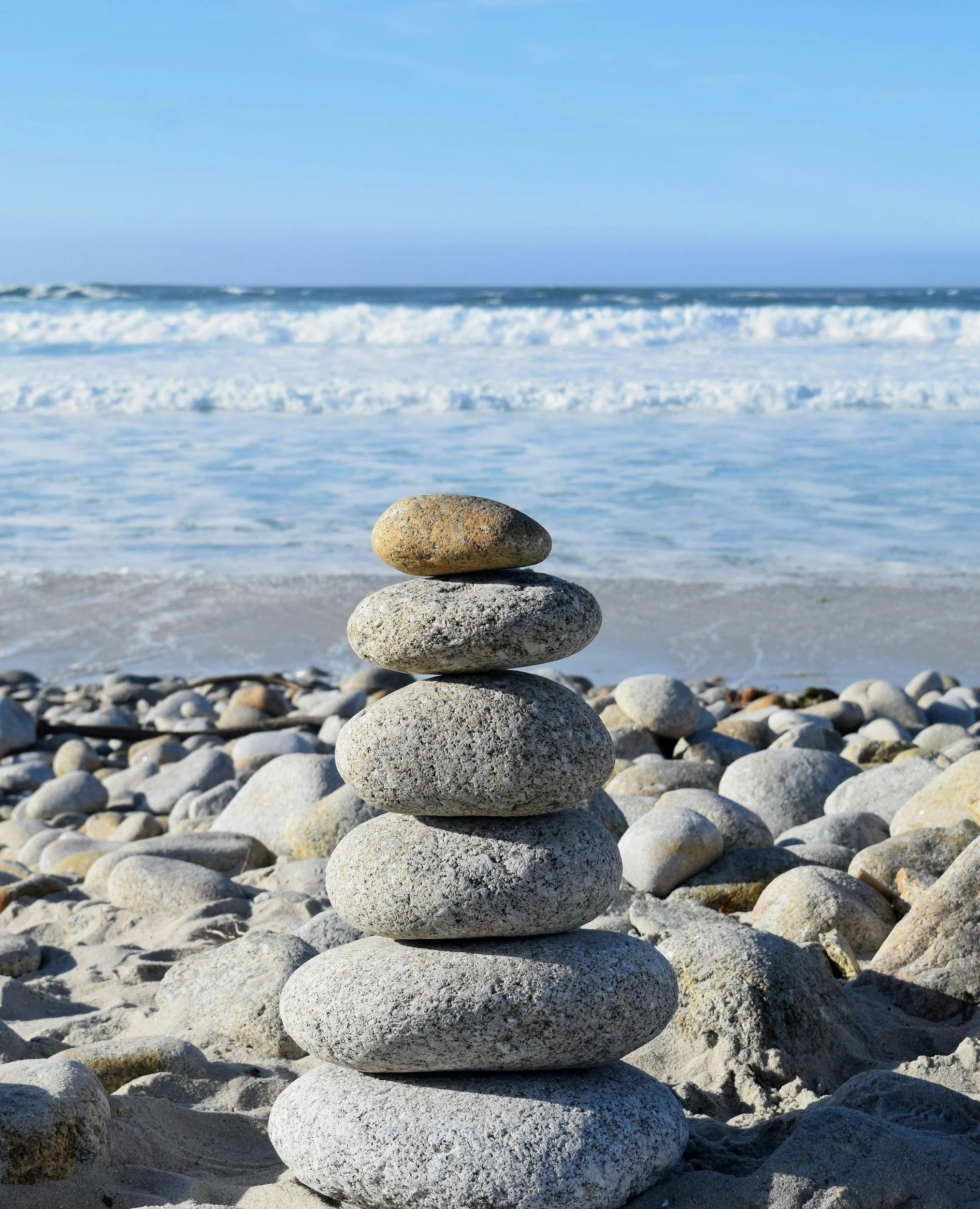 Stack of smooth stones arranged in a tower on a beach with ocean waves and blue sky in the background.
