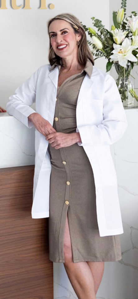 A woman with shoulder-length brown hair and light makeup standing inside a medical or office setting, wearing a beige dress with large buttons and a white lab coat, smiling at the camera, with a vase of white lilies and greenery in the background.