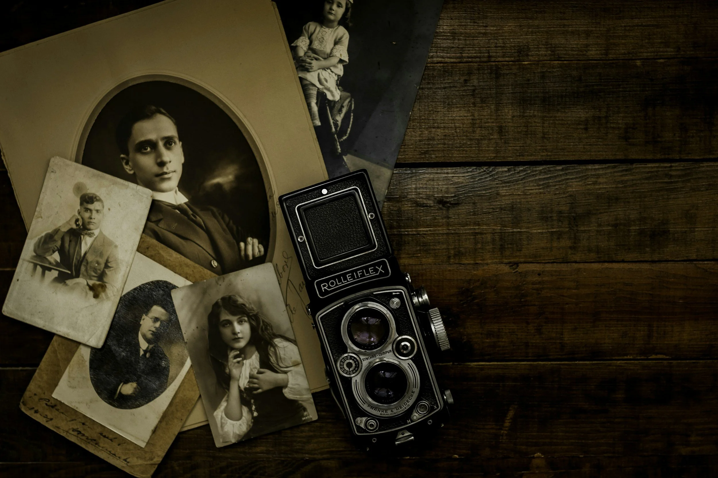 A black and white vintage camera is placed on a wooden surface next to several old photographs of people, including a woman and a young girl. The photographs are partly overlapping and have an antique look.