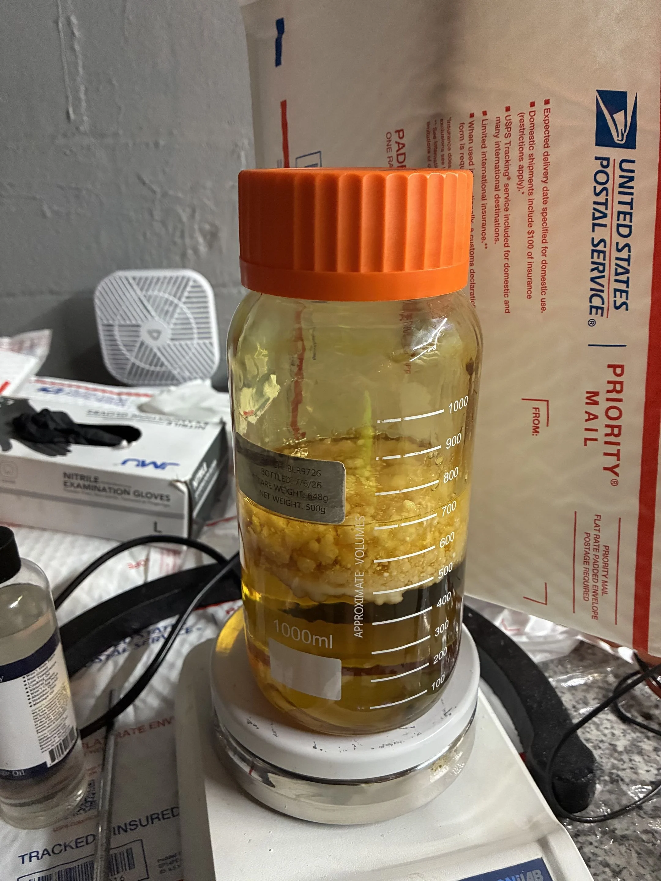 A laboratory scene with a beaker filled with a yellowish liquid and solid particles, placed on a digital scale. There are pharmaceutical or chemical bottles and boxes, including nitrile examination gloves, in the background.