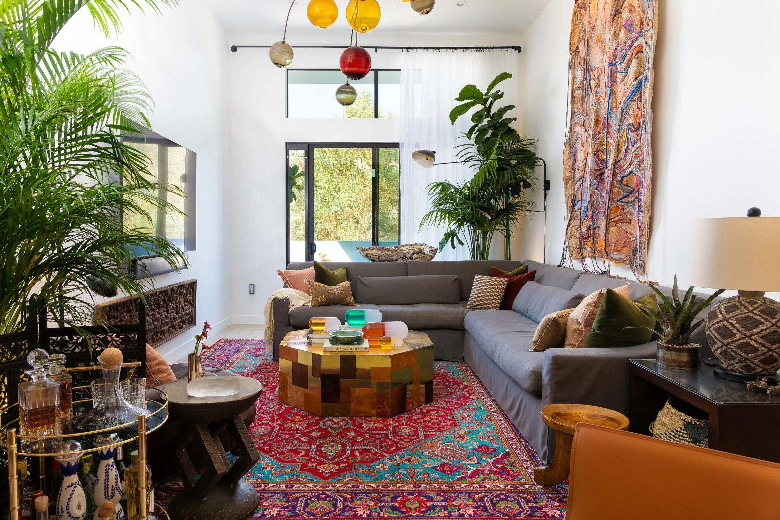 Living room at Hudson House in Hollywood, with a gray sectional, a patchwork coffee table on a red patterned rug, colored glass pendant globes and tall palms