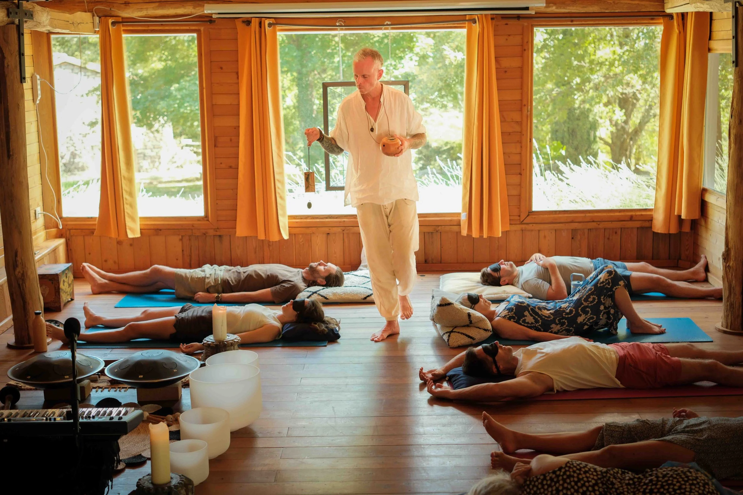 A group of people lying on their backs on yoga mats with their arms at their sides, participating in a meditation or yoga class inside a wooden room with large windows and yellow curtains. A person stands in the center, conducting the session, holding a small object. The room is decorated with candles and musical instruments, with natural greenery visible outside the windows.