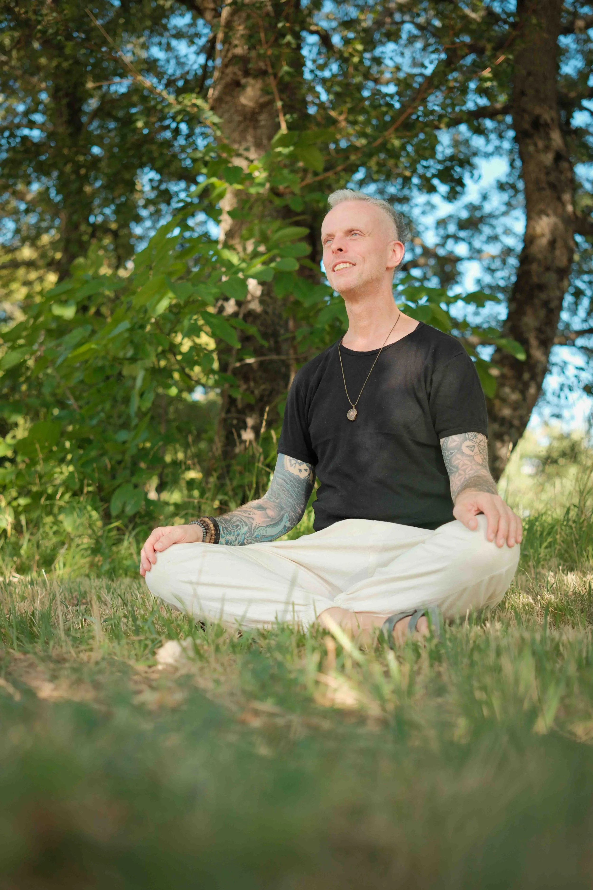 A man with tattoos, short gray hair, and a necklace, sitting cross-legged outdoors on grass, smiling with eyes closed in front of a large tree and blue sky.