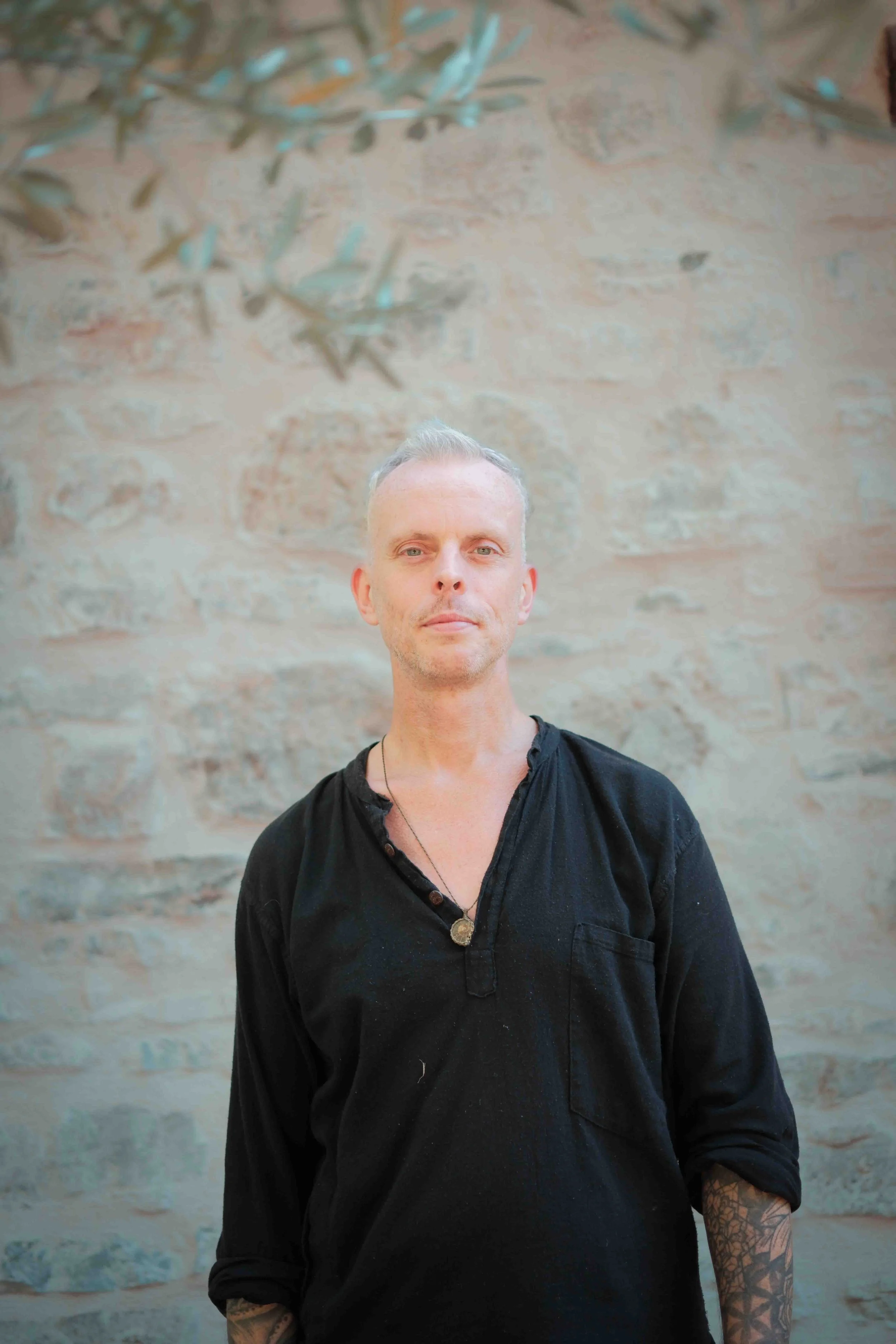 A man with short gray hair and tattoos on his arms standing outdoors in front of a stone wall.