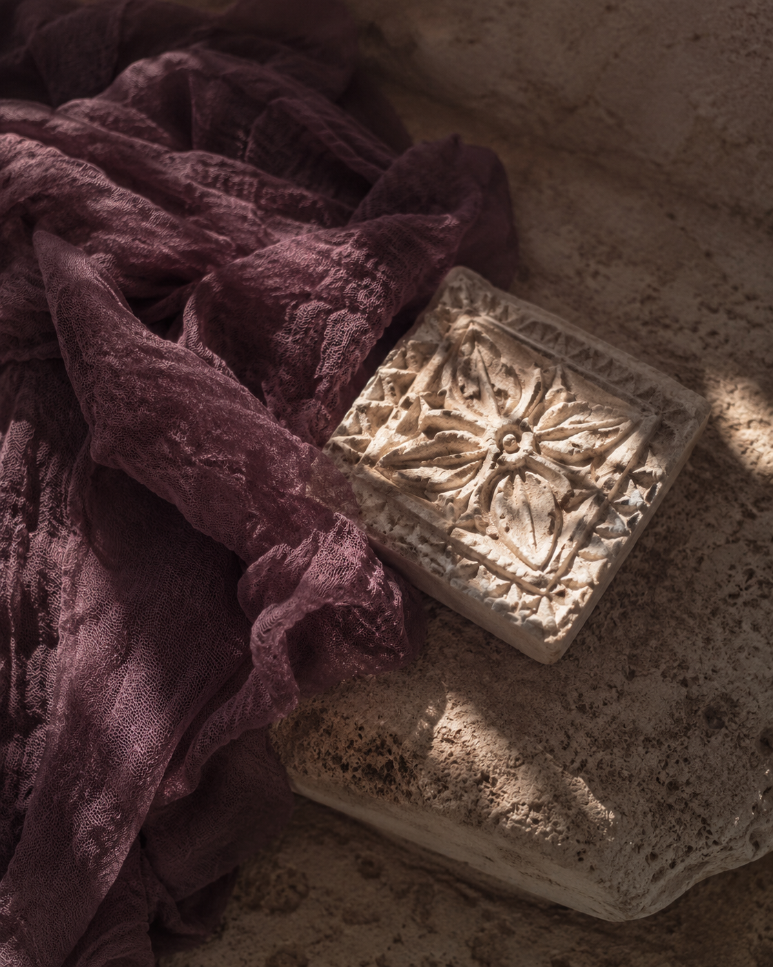 A decorative stone tile with an intricate floral pattern next to a wrinkled purple cloth, on a textured stone surface.