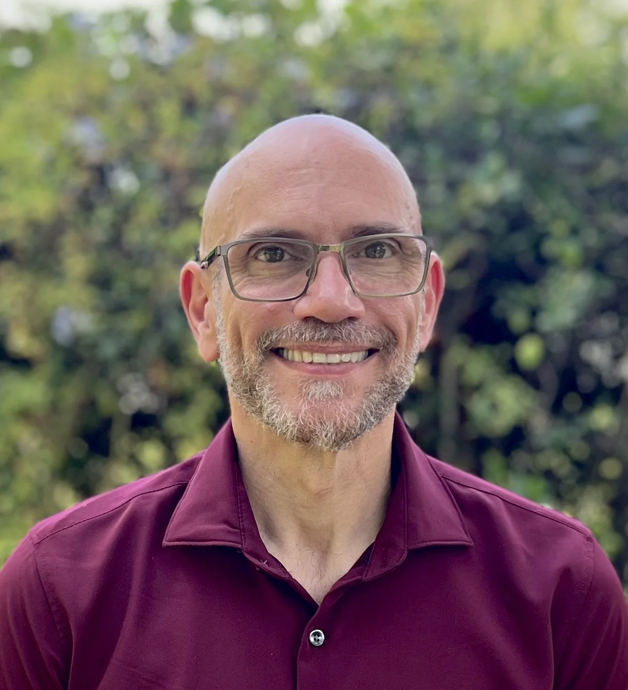 A smiling bald man with glasses, a gray beard, and a mustache, wearing a maroon shirt, standing outdoors with green trees in the background.