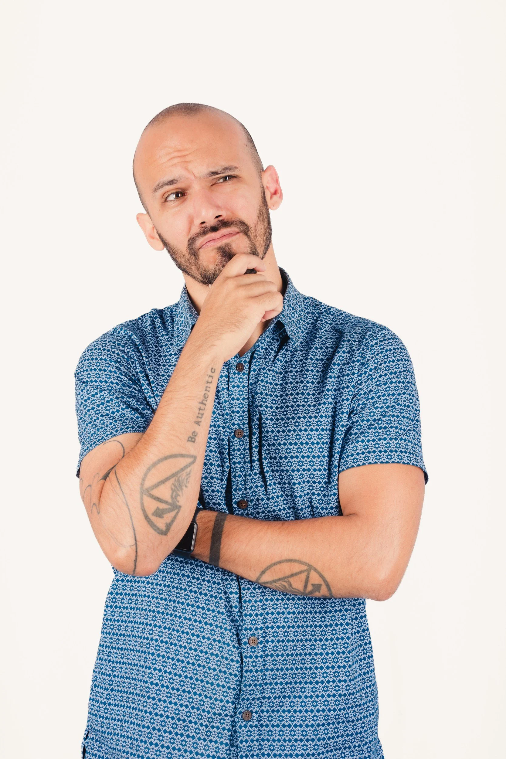 A man with a shaved head and beard, wearing a blue patterned short-sleeve shirt, is thinking with his finger on his chin and looking to the side.