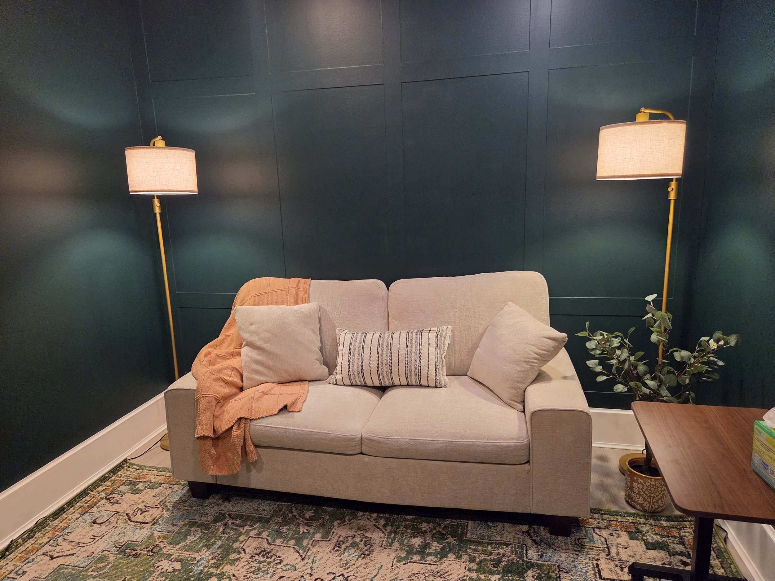 Living room corner with off-white sofa, orange blanket draped over arm, striped throw pillow, dark teal wall, two yellow floor lamps with fabric shades, green potted plant, wooden side table, patterned area rug.