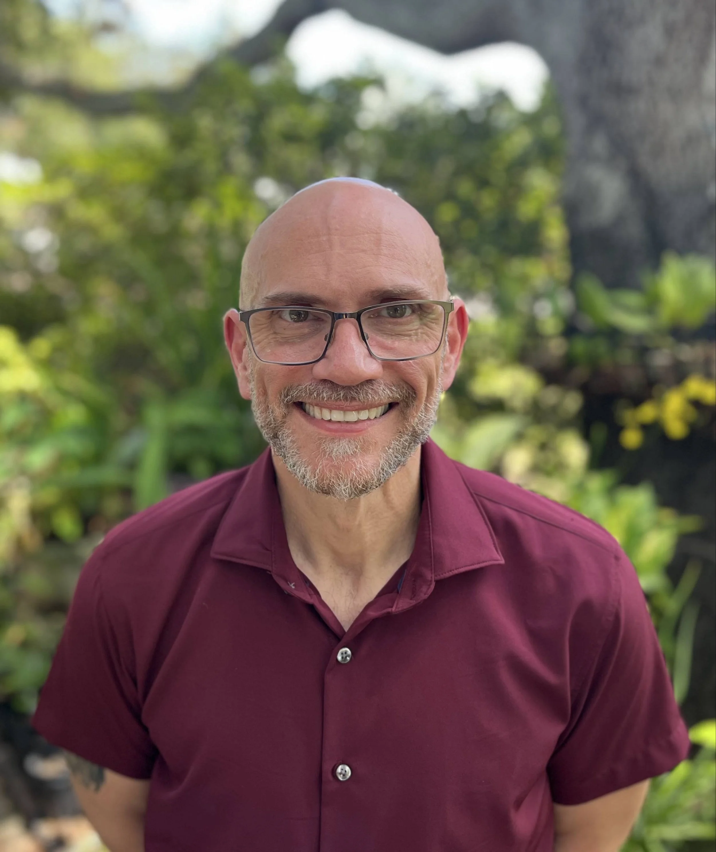 Close-up of a smiling man with glasses, a shaved head, gray beard, wearing a maroon shirt, outdoors with green foliage in the background.